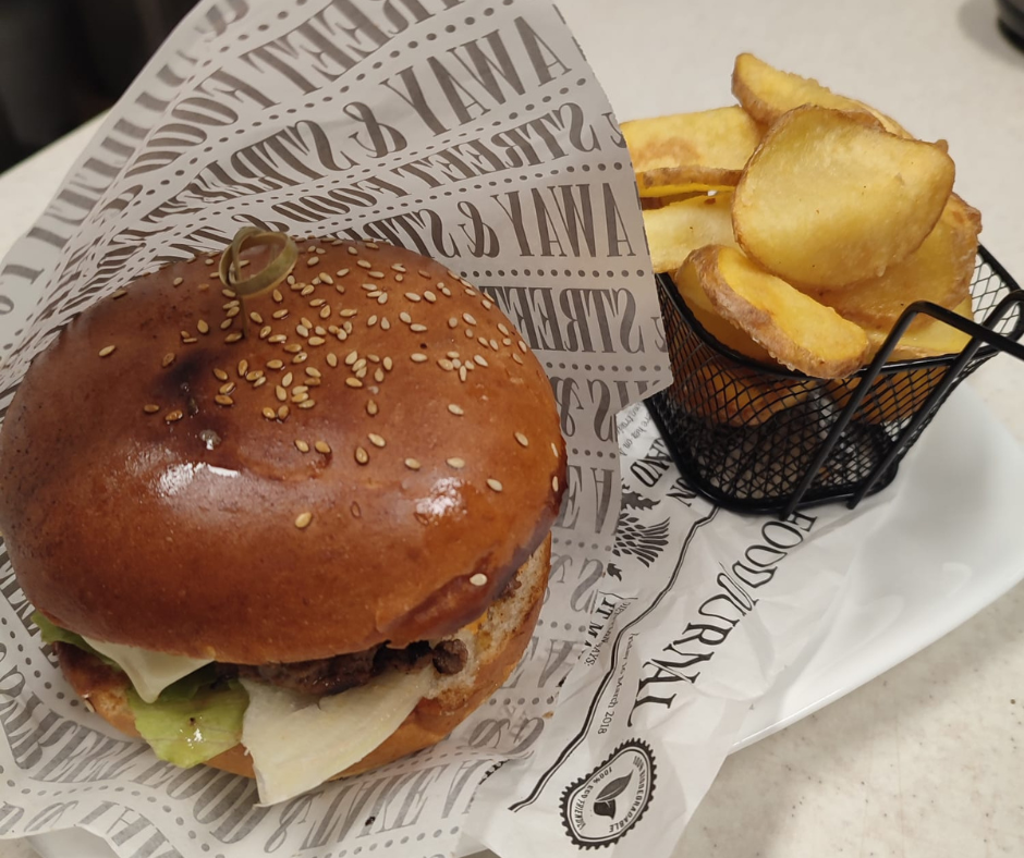 Hamburger with lettuce and cheese in a sesame seed bun, served with a side of thick-cut potato fries in a small black wire basket.