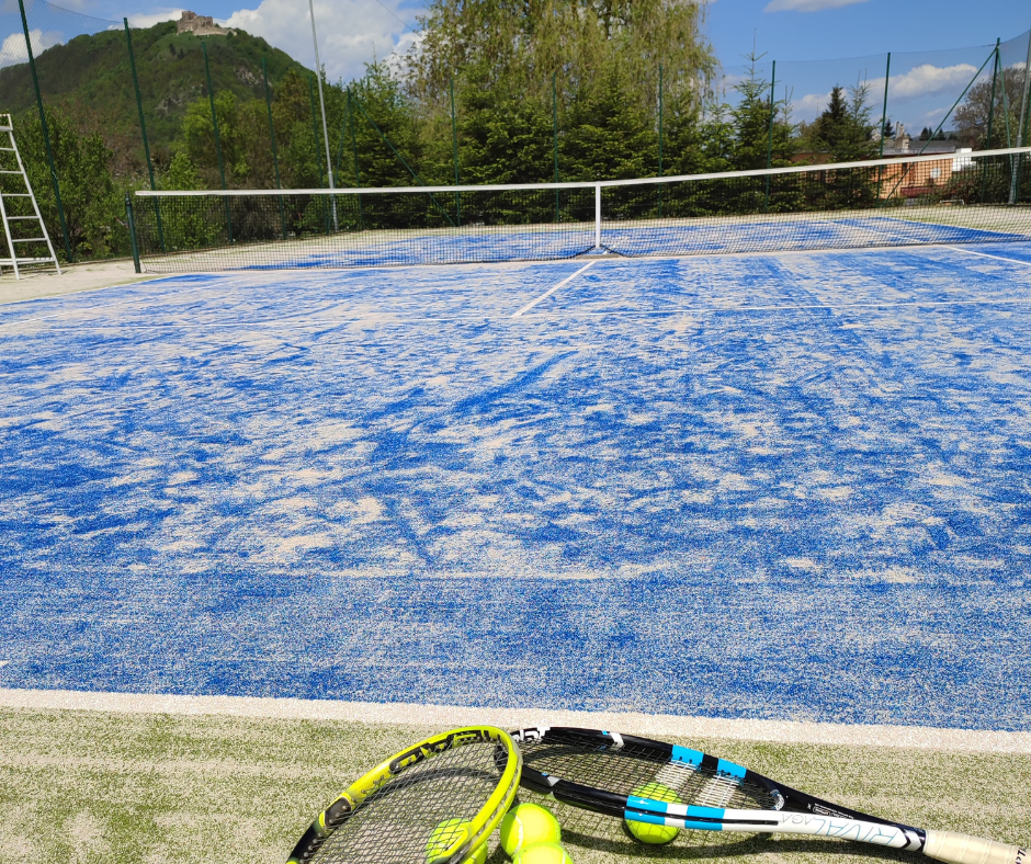 Empty outdoor tennis court with sand-covered surface, surrounded by a green fence, with tennis rackets and balls in the foreground and trees and a hill with a castle in the background.