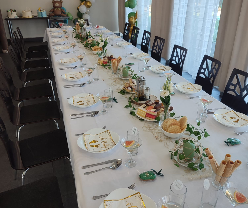 A long dining table set for a celebration with white tablecloth, decorative plates, glasses, and silverware. The table is adorned with flowers, greenery, and assorted desserts. In the background, balloons and a teddy bear decoration are visible, indi