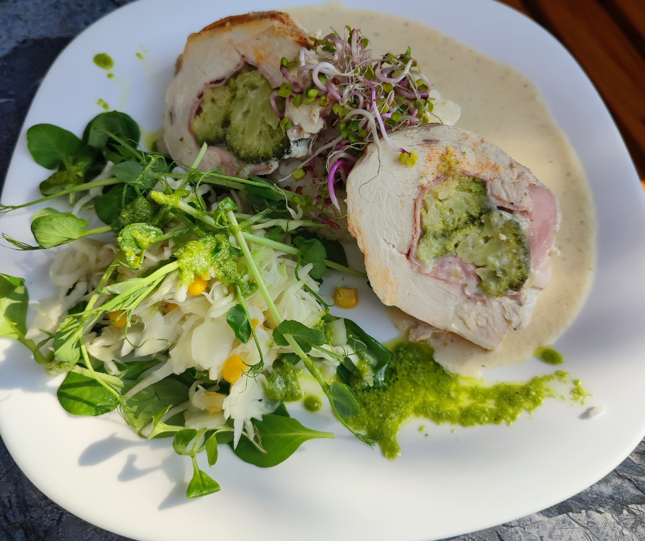 Stuffed chicken breast with green sauce served with a side salad topped with microgreens and shredded vegetables on a white plate.