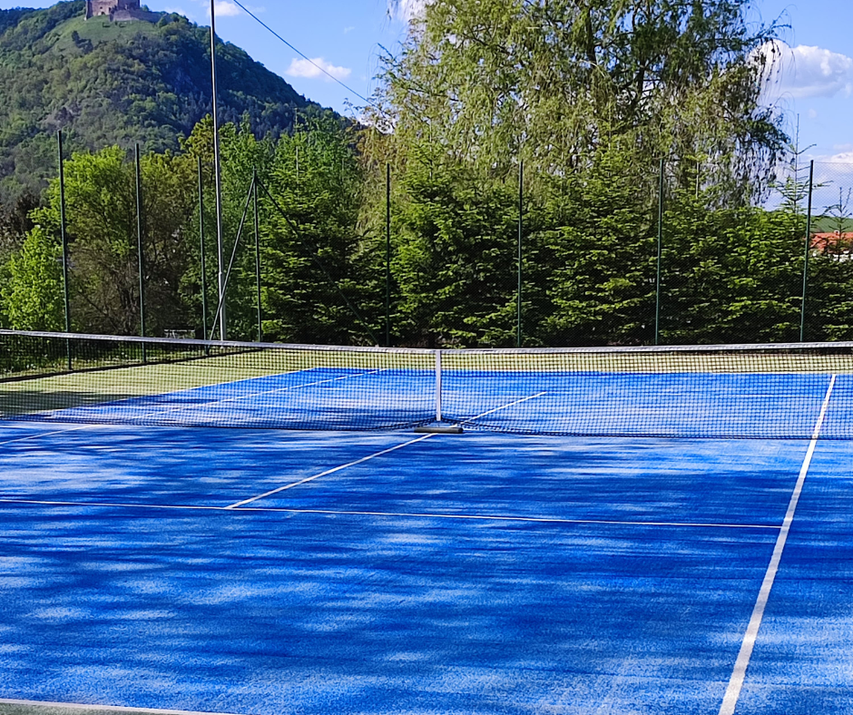 An outdoor tennis court with a blue playing surface, surrounded by trees and a mountain in the background, under a partly cloudy sky.
