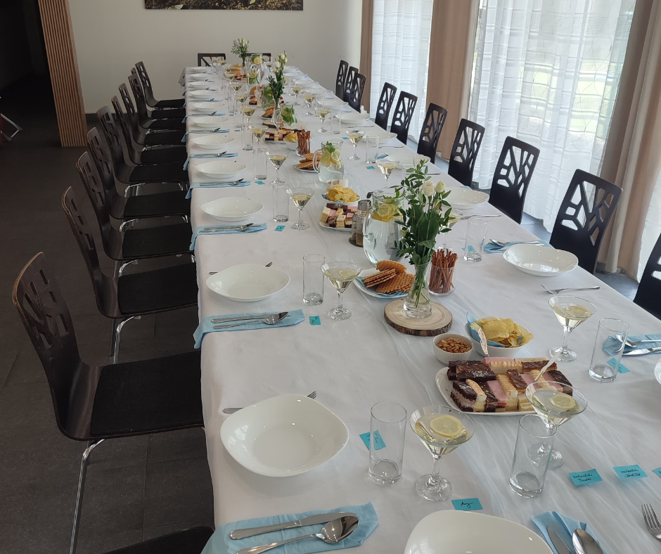 Long dining table set for a formal meal with plates, glasses, cutlery, and centerpieces, along with various appetizers and desserts, in a well-lit room with large windows and curtains.