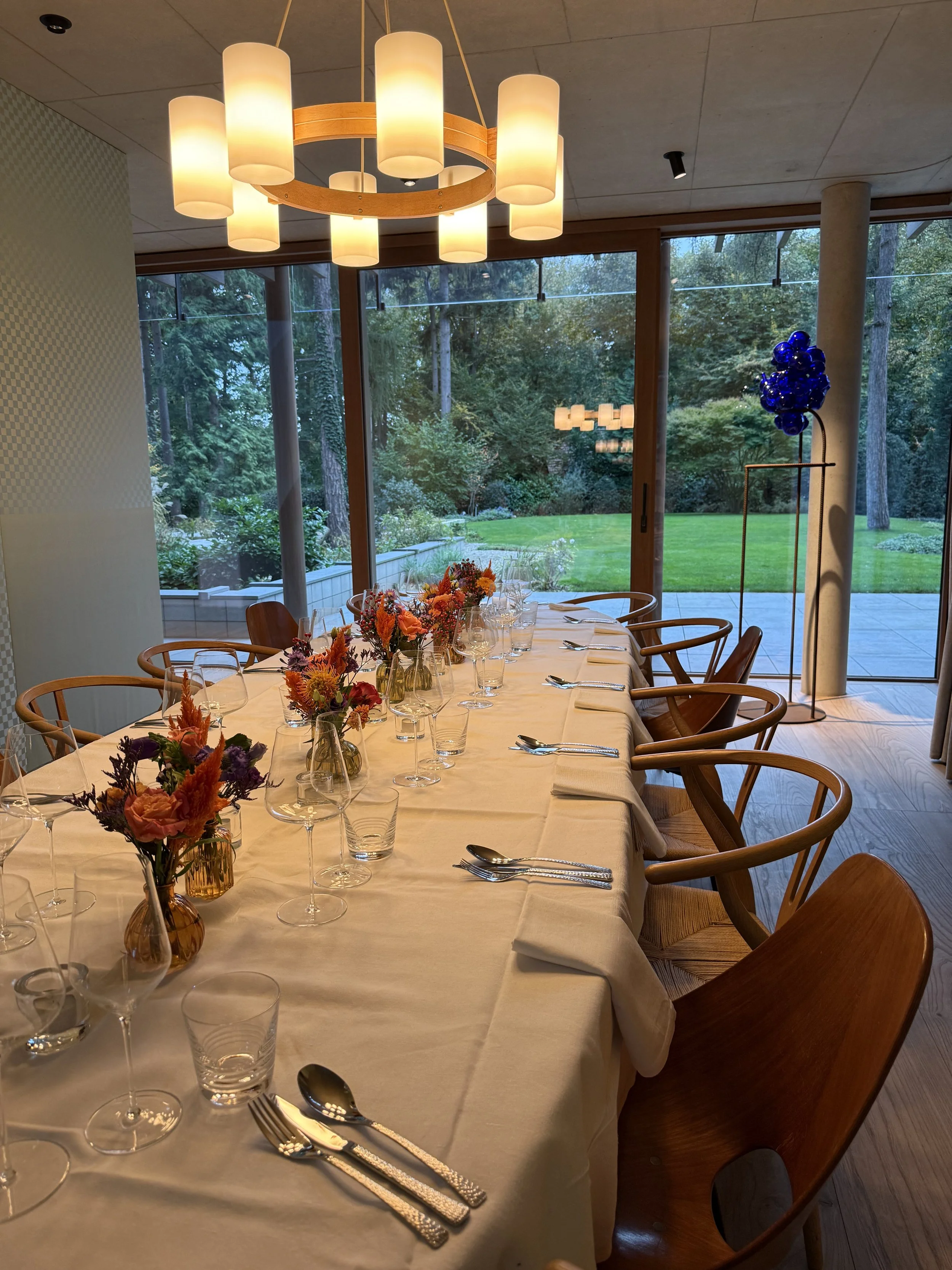 Een lange diningtafel afgelegd met een witte tafelkleed, versierd met bloemen in kleine vaasjes, glazen en bestek. De kamer heeft grote ramen die uitkijken op een groen buitengebied, met een moderne plafondlamp erboven. #chefvandenberghe , chef van d