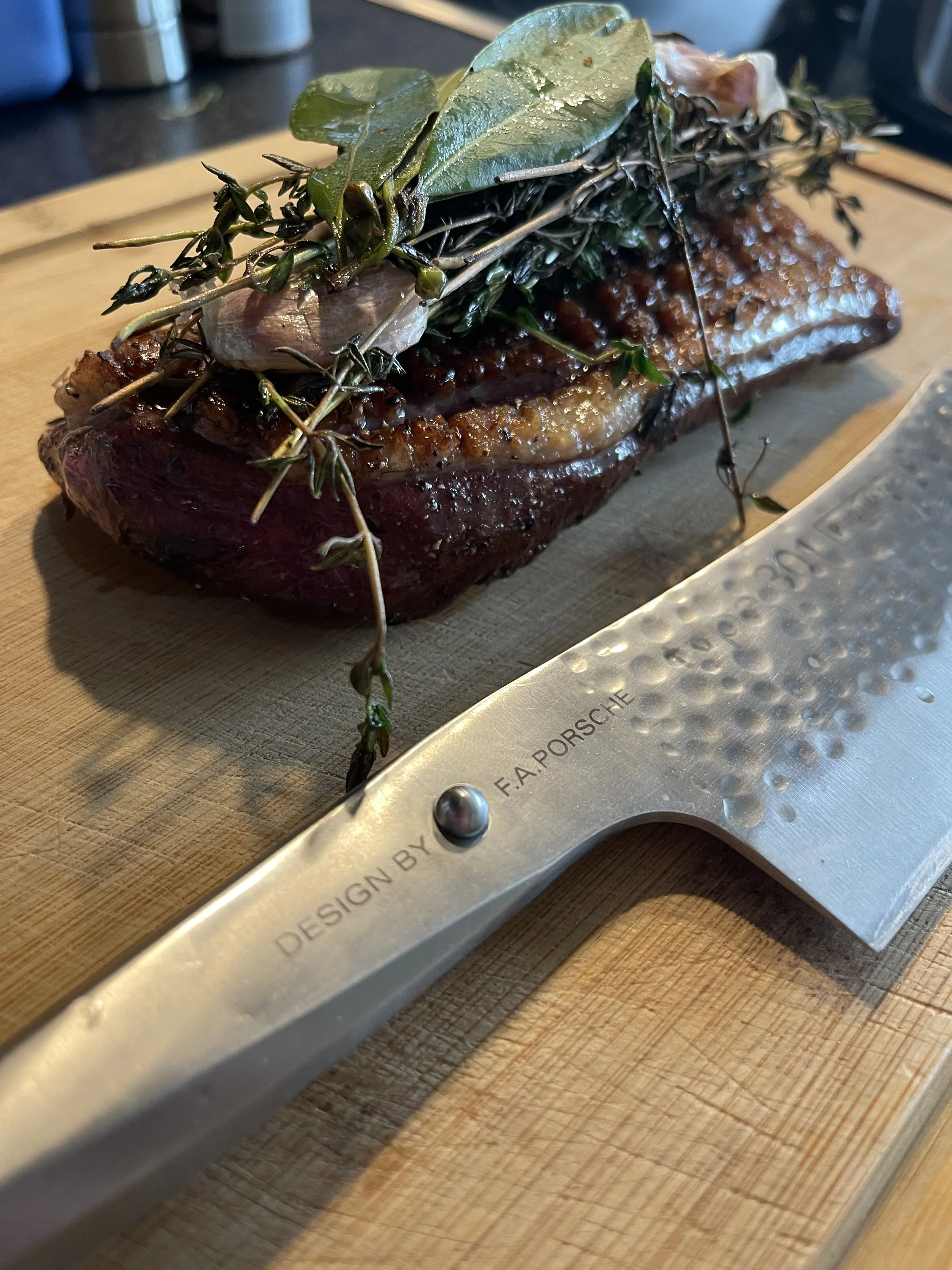 Gekookt stuk vlees met kruiden en kruiden op een houten snijplank, met een geribbelde messcherp aan de rand.