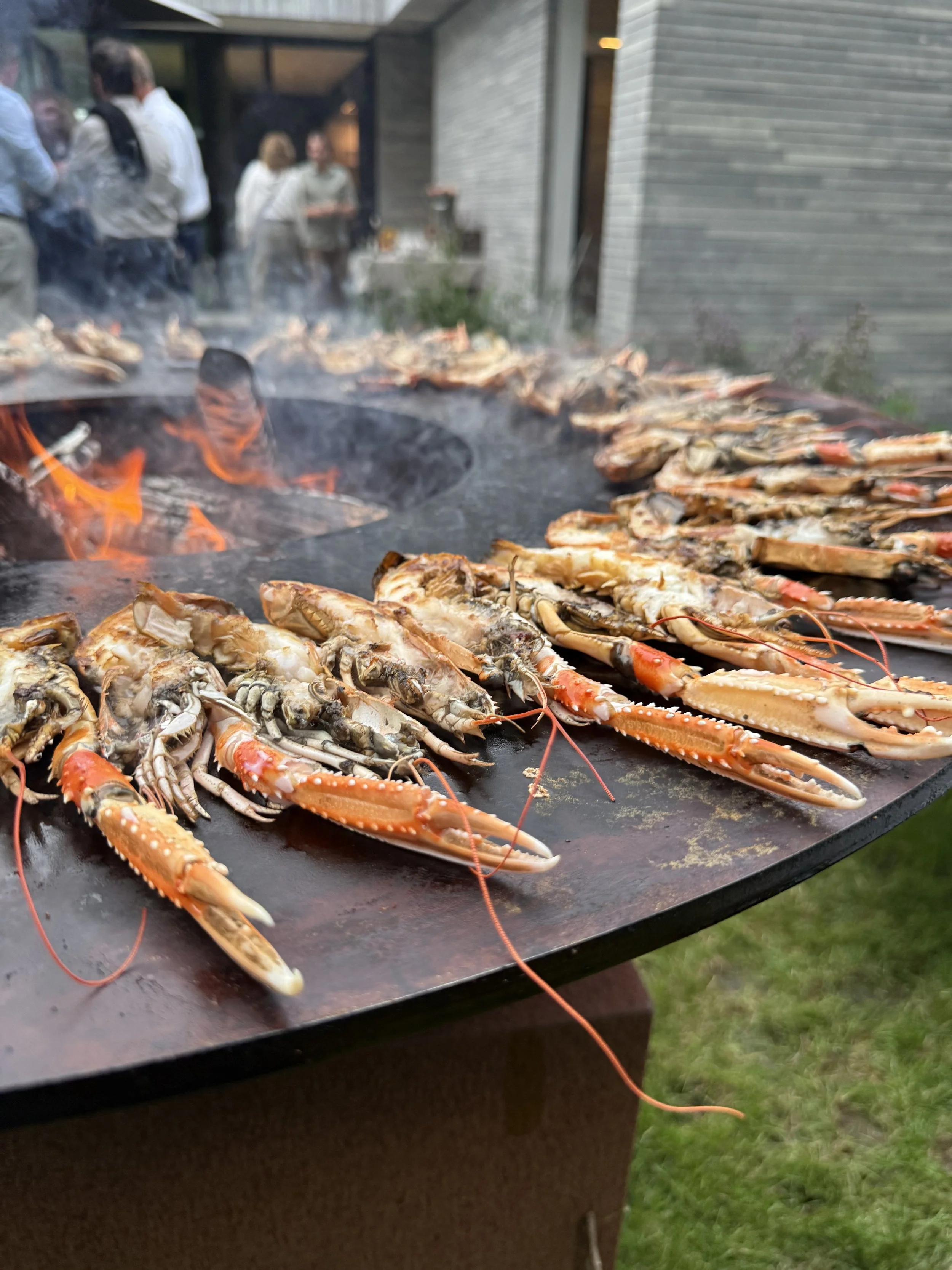 Gerookte kreeften op een barbecue met een groep mensen op de achtergrond. #chefvandenberghe , chef van den berghe , private dining, private chef aan huis