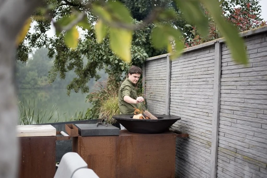 Een man die buiten aan het barbecueën is naast een houten hek met water en bomen op de achtergrond. #chefvandenberghe , chef van den berghe , private dining, private chef aan huis