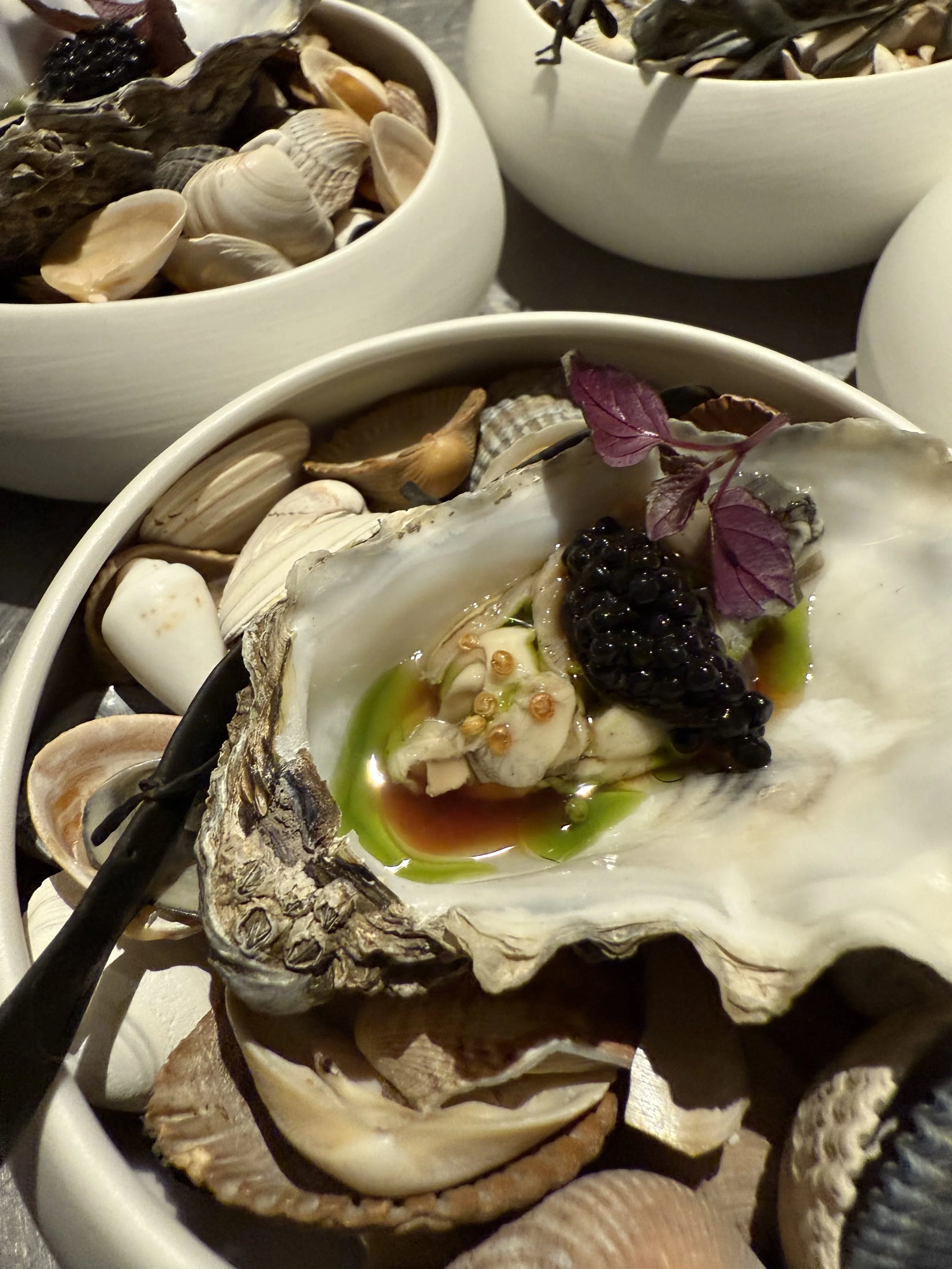 Close-up van een oester met zwarte kaviaar, groene olie, saus, een paar blaadjes purple basilicum en schelpen in witte kommen en schaaltjes op een donkere tafel. #chefvandenberghe , chef van den berghe , private dining, private chef aan huis