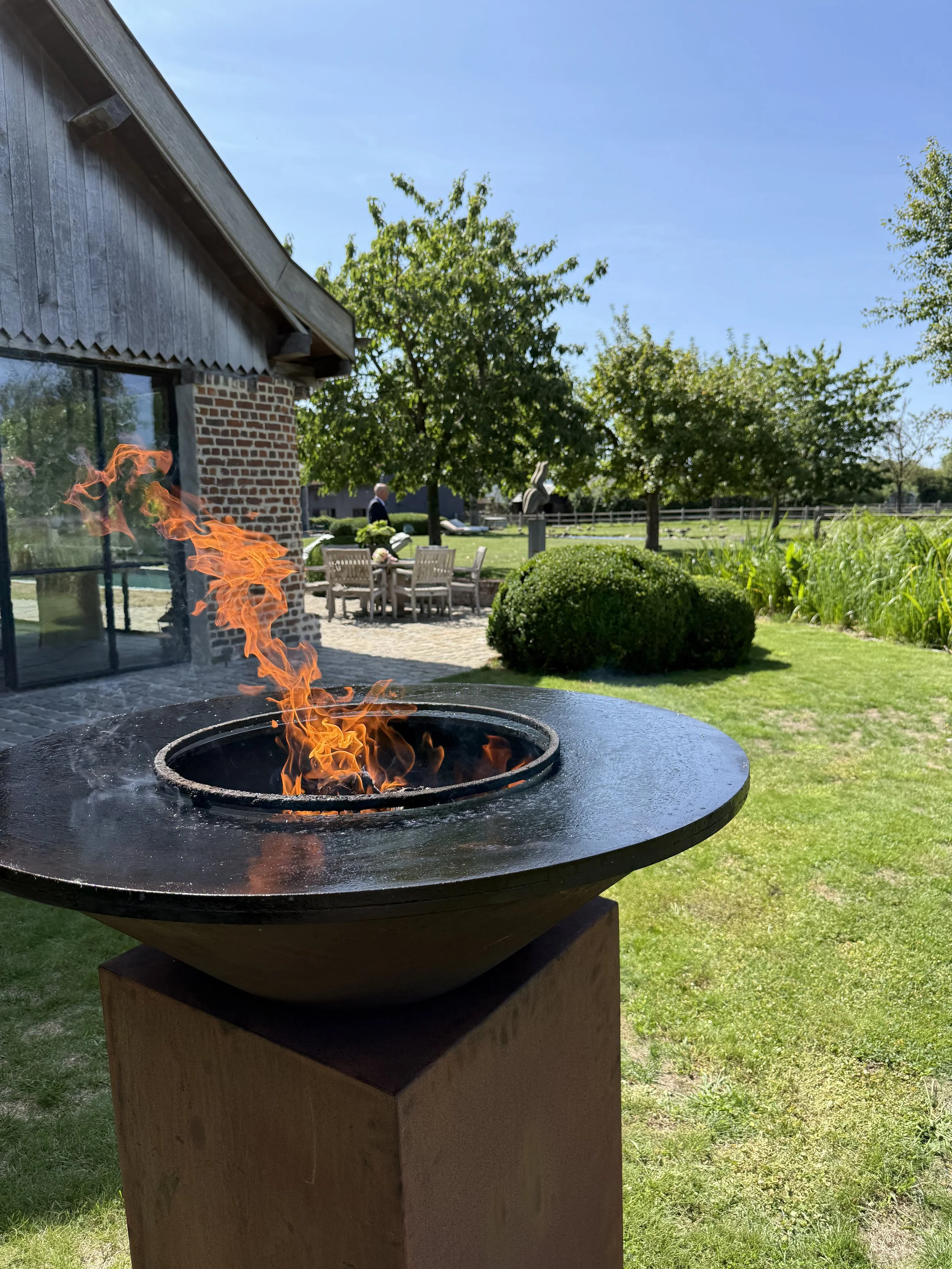 Een buitengrill met vlammen tegen een zonnige dag met groene bomen, gras, struiken en een huis op de achtergrond. #chefvandenberghe , chef van den berghe , private dining, private chef aan huis