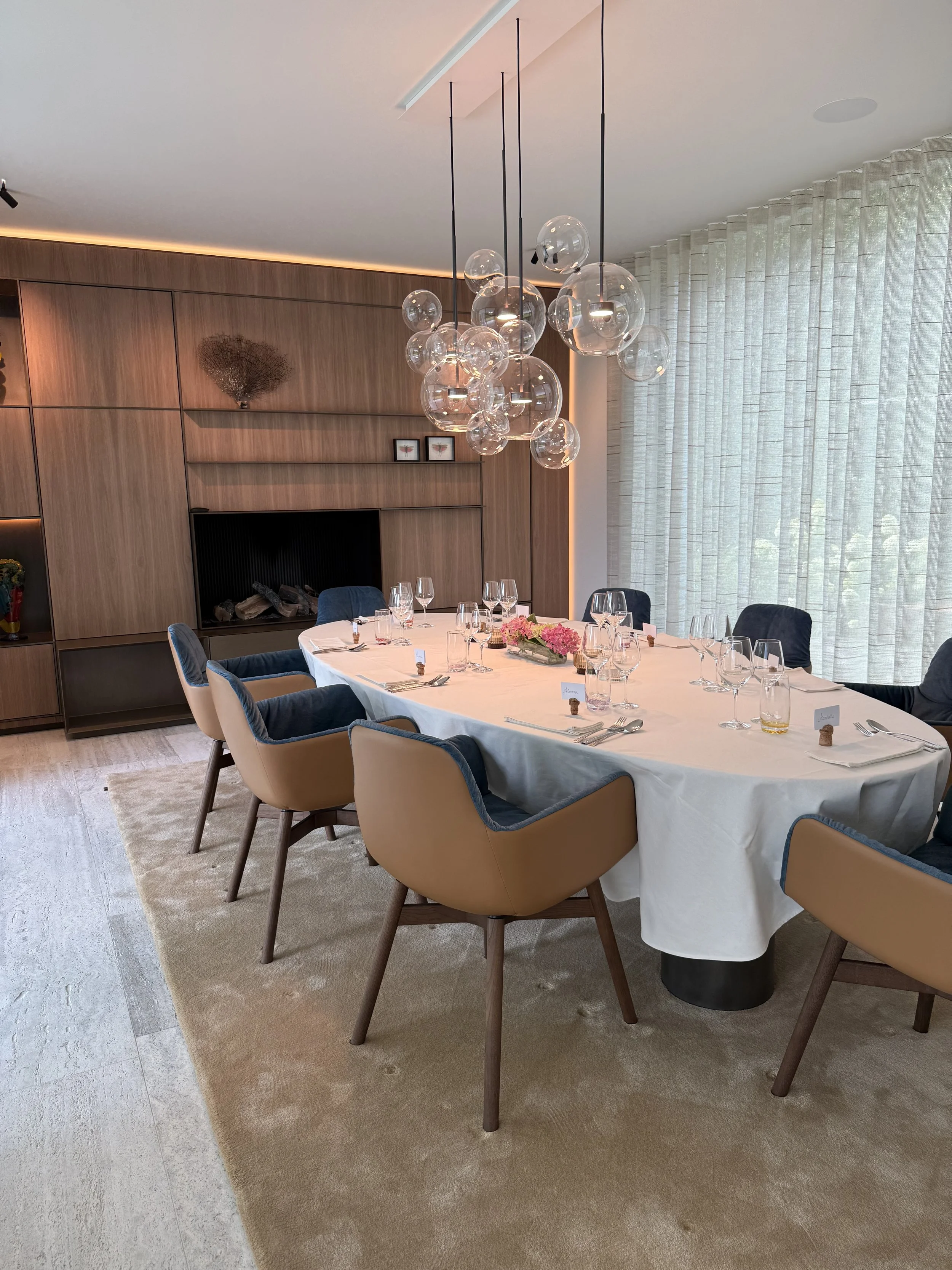 Een elegante eettafel met een witte tafelkleed, versierd met glazen, bestek, en een bloemenarrangement in het midden. Er zitten zes empty stoelen rondom de tafel, drie aan elke zijde. De kamer heeft grote ramen met witte gordijnen en een warme houten wand met decoraties, inclusief een abstracte houten muurkunst en een open haard. Een moderne glazen hanglamp hangt boven de tafel.