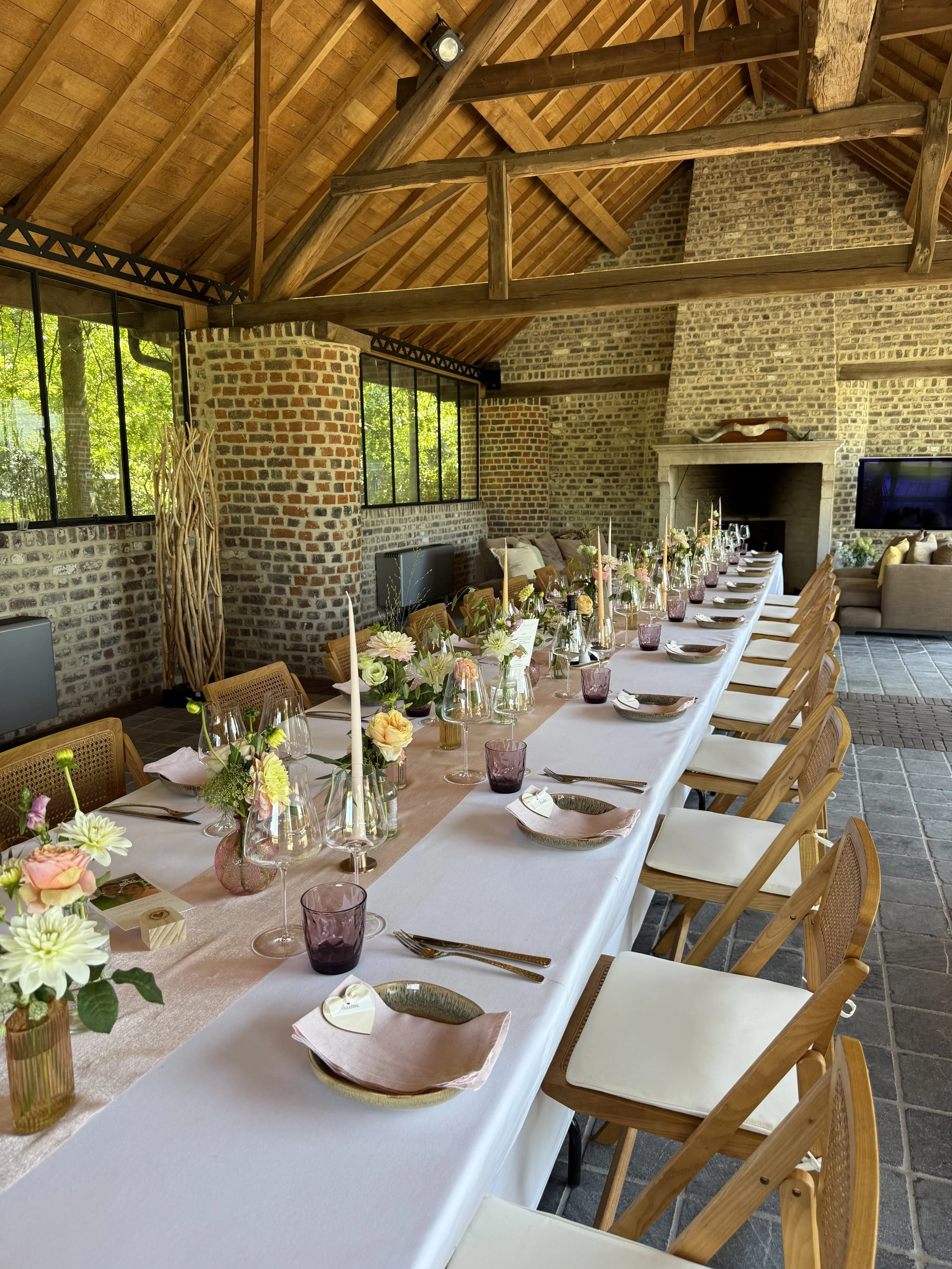 Een lange eettafel met bloemen, glazen en borden, klaar voor een etentje in een rustieke kamer met stenen muren en houten plafond. #chefvandenberghe , chef van den berghe , private dining, private chef aan huis