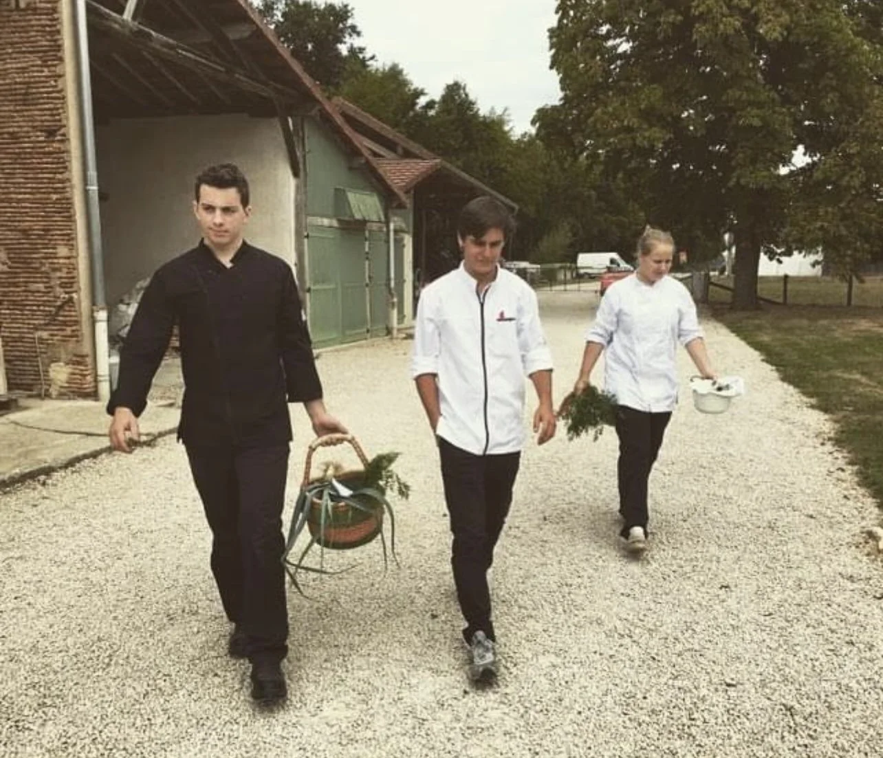Drie mensen lopen op een grindpad in een landelijke omgeving, twee dragen manden met groenten en één draagt een metalen pan, allemaal zijn gekleed in witte of zwarte kledij, en er zijn bomen en gebouwen op de achtergrond. #chefvandenberghe , chef van