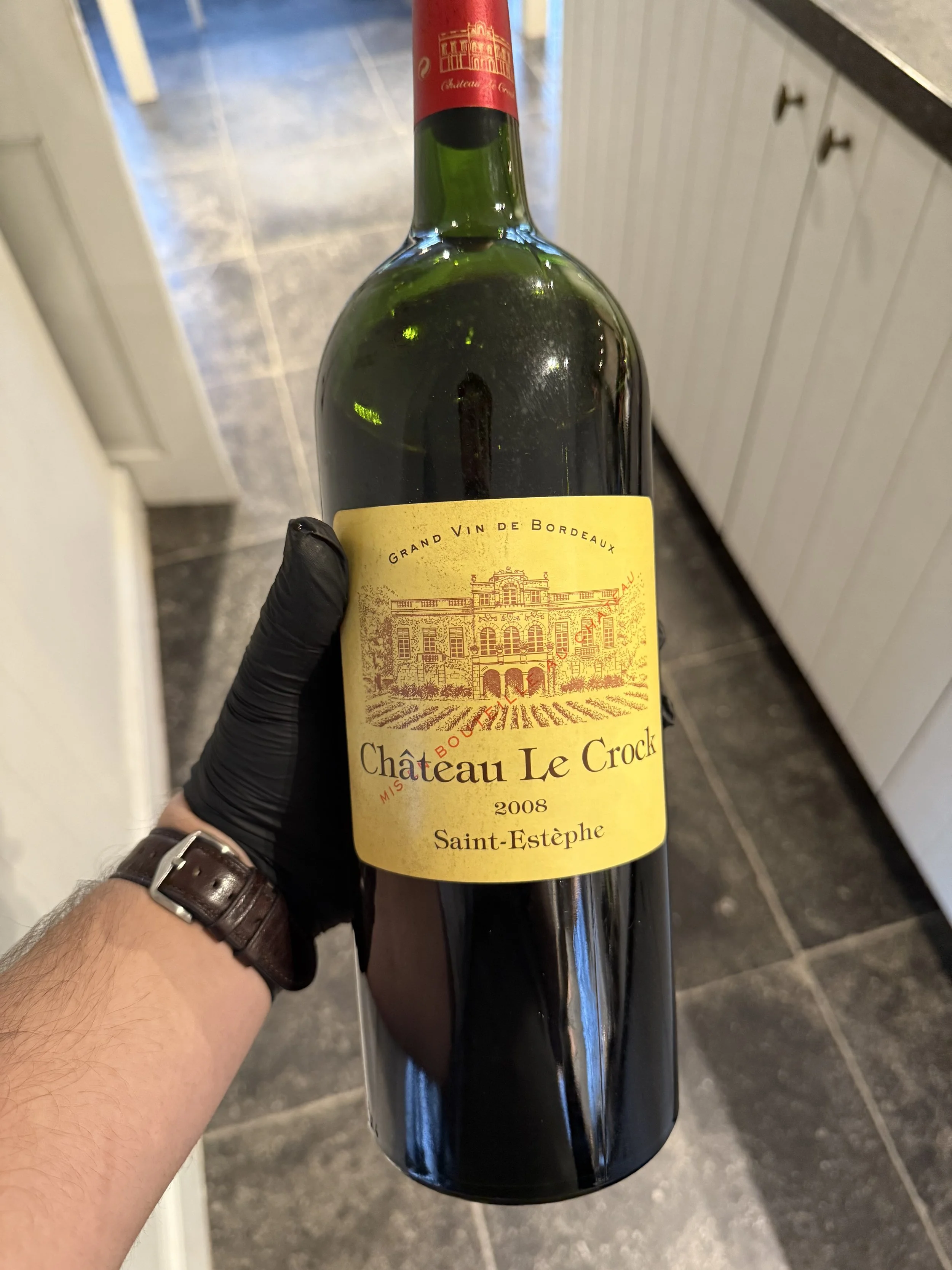 Een hand die een fles rode wijn vasthoudt, met een geel wijnetiket dat 'Château Le Crock 2008 Saint-Estèphe' en een afbeelding van een kasteel weergeeft. #chefvandenberghe , chef van den berghe , private dining, private chef aan huis