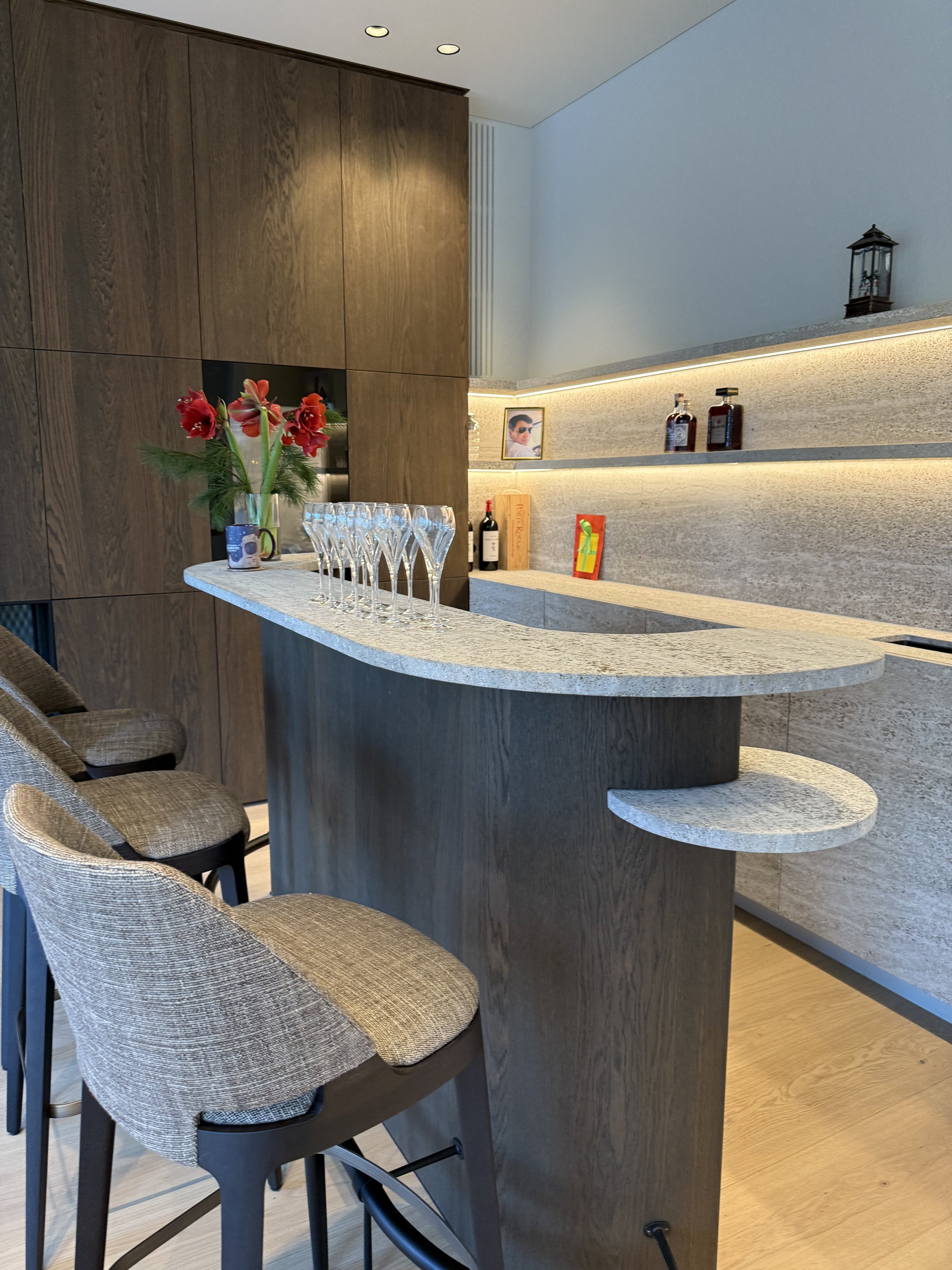 Een moderne keuken met een ontbijtbar met drie barkrukken, bloemen in een vaas, en decoratieve items op de planken. #chefvandenberghe , chef van den berghe , private dining, private chef aan huis