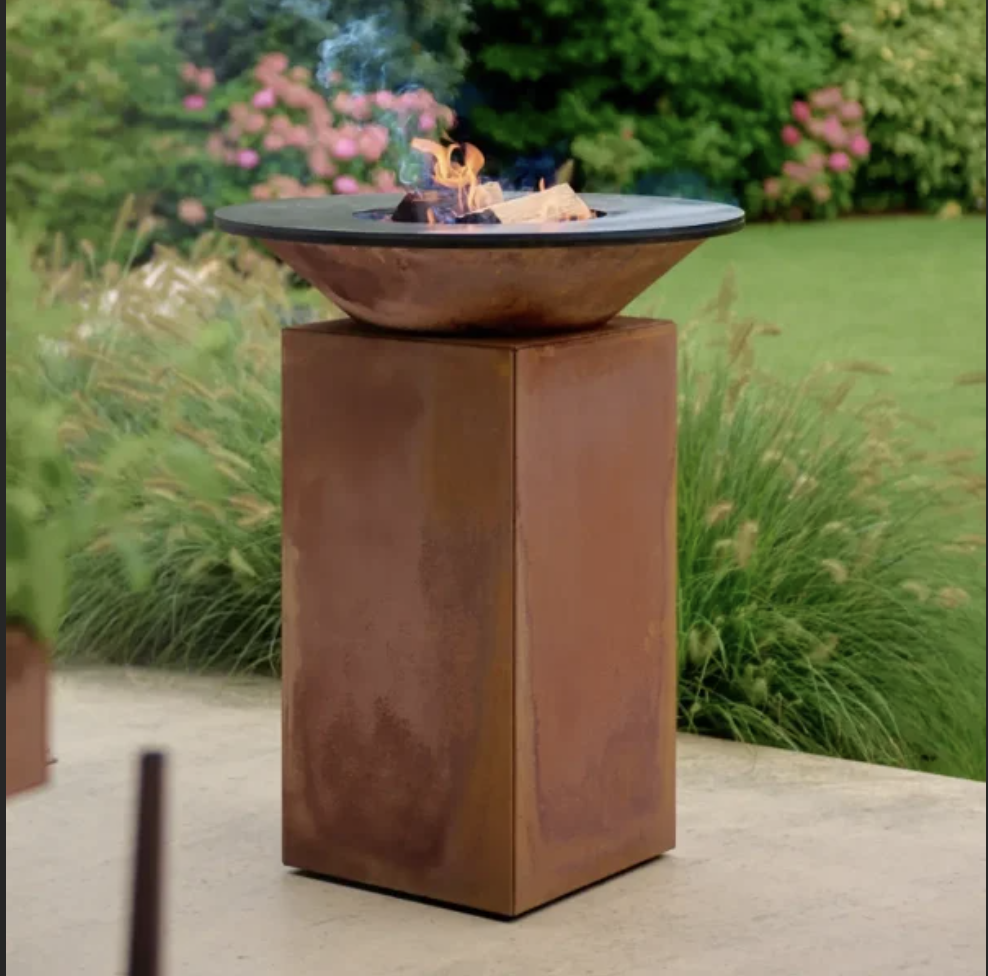 Een metalen schaal met brandend hout op een roestkleurige sokkel, zich geheel in een tuin met groen gras en bloemrijke struiken. #chefvandenberghe , chef van den berghe , private dining, private chef aan huis