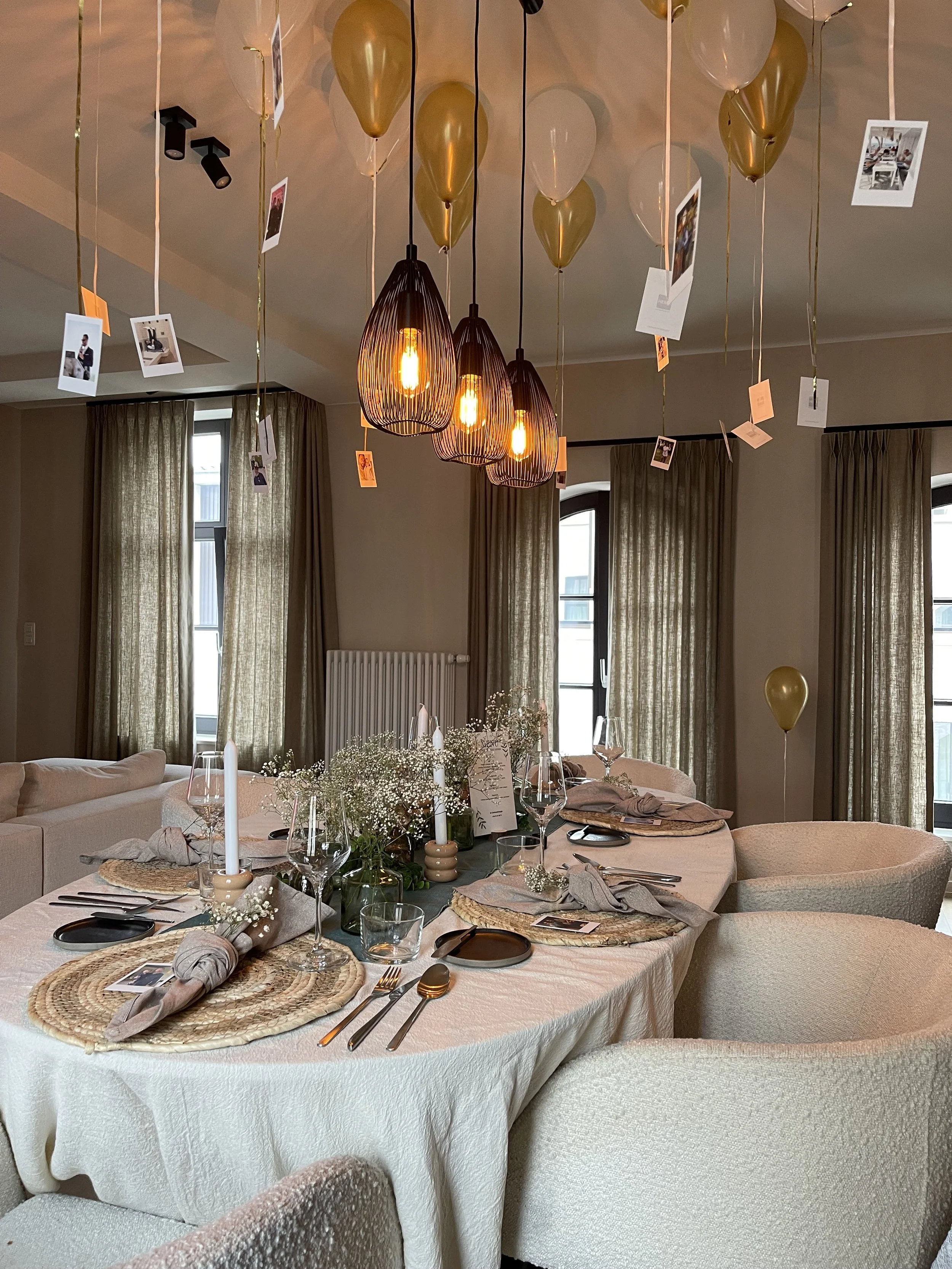 Een elegante feesttafel met witte tafelkleed, versierd met bloemen, kaarsen, bestek, glazen en placemats, in een sfeervol ingeholde kamer met beige gordijnen en een verlichte hanglamp met ballonnen en Polaroid foto's boven de tafel. #chefvandenberghe