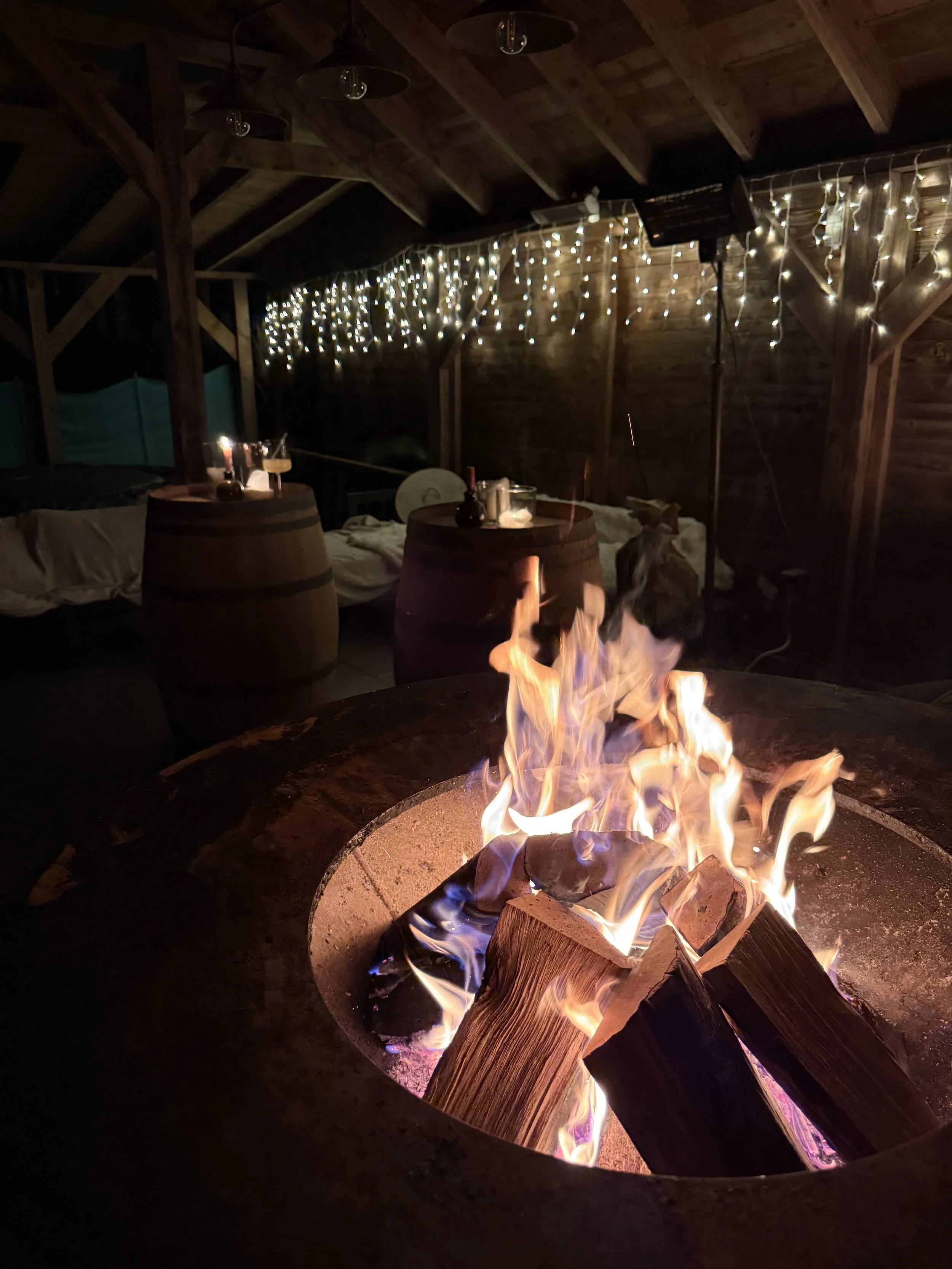 Knusvuur met brandende houtblokken in een gezellige, sfeervolle ruimte met houten wanden en plafond, versierd met witte stringlichten. #chefvandenberghe , chef van den berghe , private dining, private chef aan huis