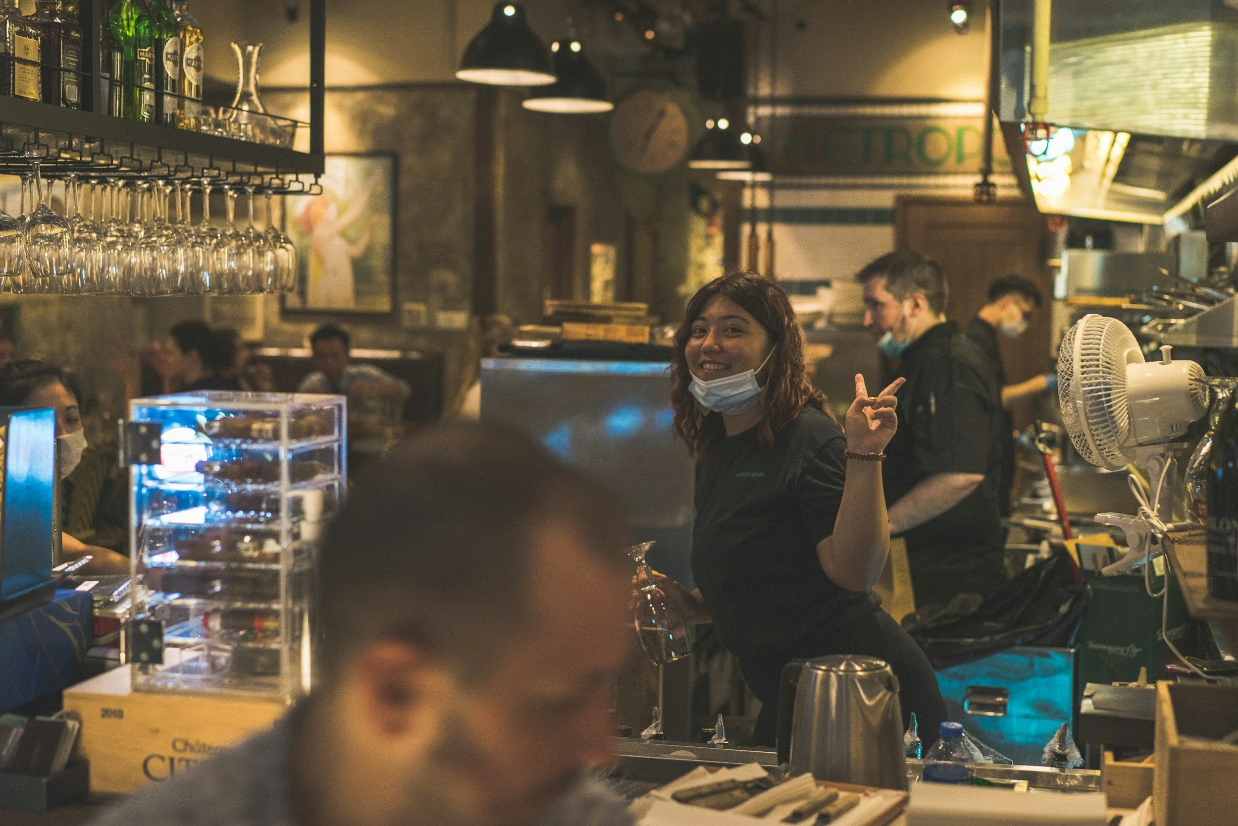 A smiling waitress in a black uniform and face mask pointing at the camera inside a busy restaurant or bar with other staff and customers in the background.