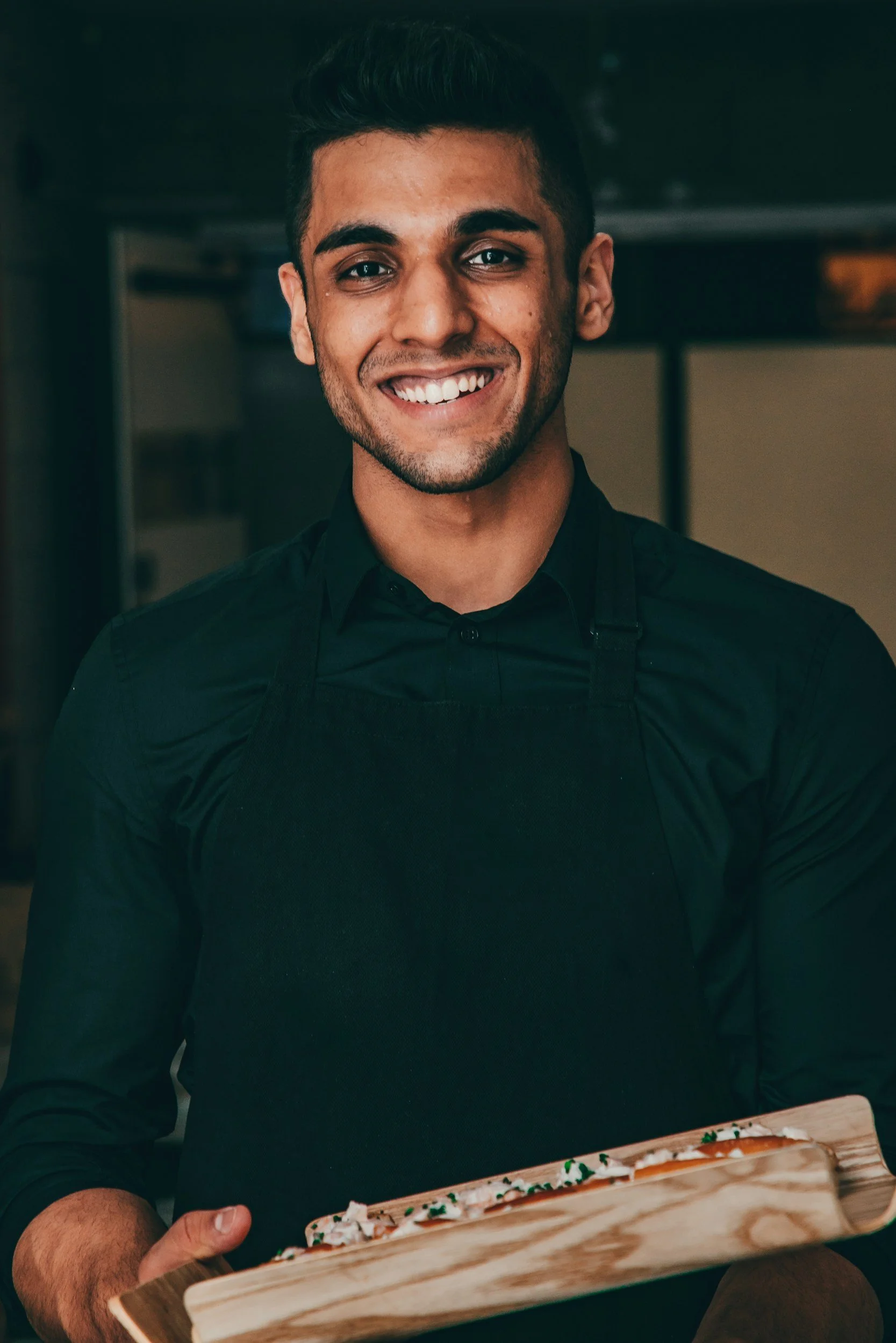 A young man smiling and holding a wooden tray with what appears to be a decorated pizza or flatbread, in an indoor setting.