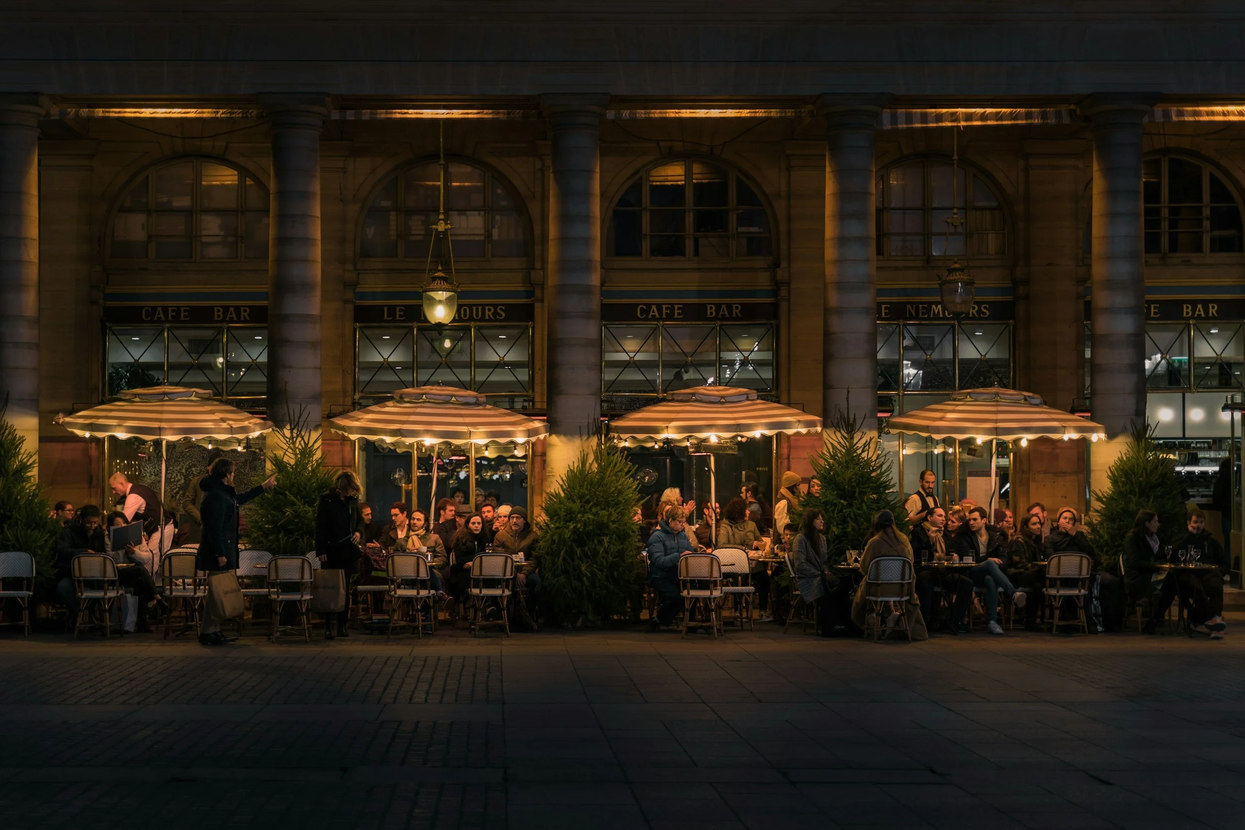 People dining outside at a cafe restaurant in the evening, with large umbrellas and potted plants, in front of a historic building with arched windows and columns.