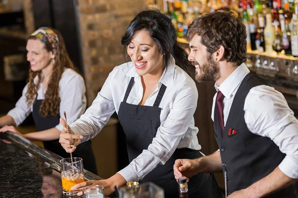 Two bartenders, a woman and a man, are smiling and preparing drinks behind a bar with liquor bottles, while another woman in the background is working.