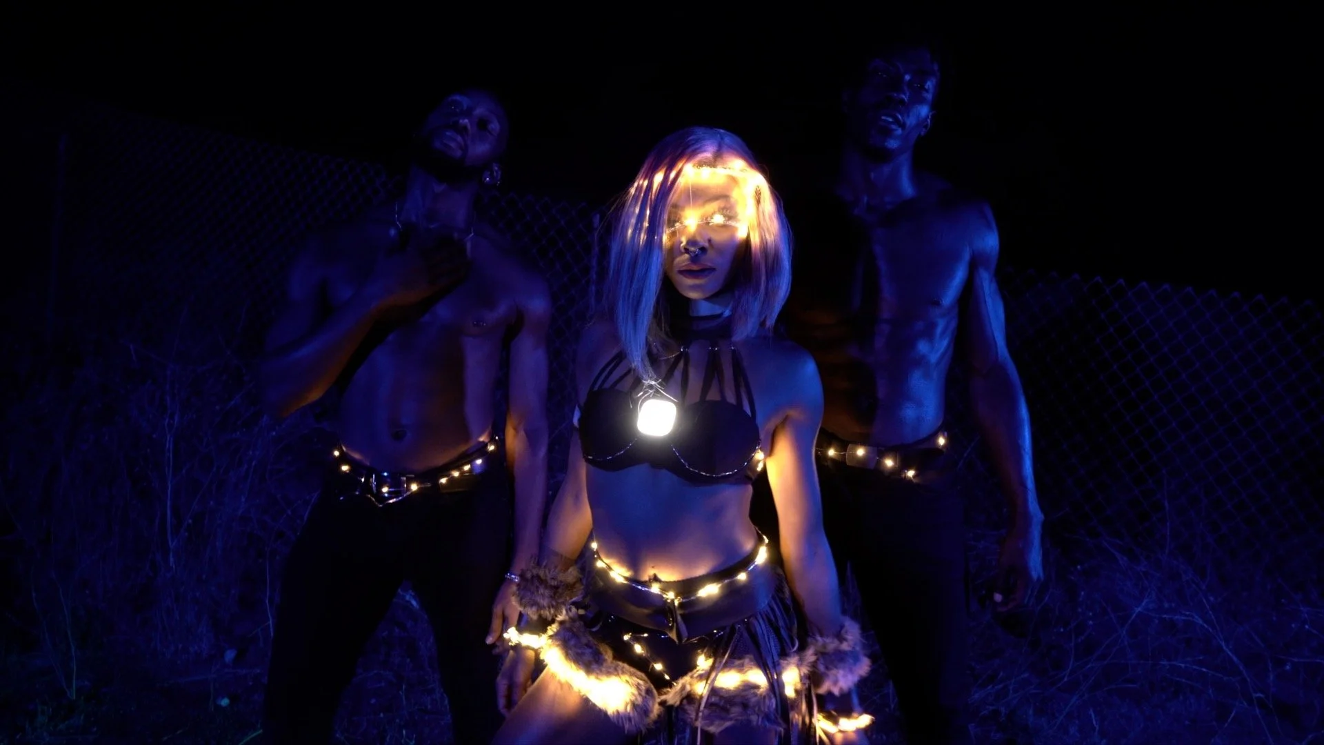 Three individuals dressed in dark, illuminated costumes at night. The woman in the center has shoulder-length blonde hair, glowing glasses, and a shining necklace, with her hand on her thigh. The two men on either side are shirtless or wearing minimal clothing, with dark skin, and are standing against a dark background with a fence. The scene is dark with blue and yellow lighting effects.