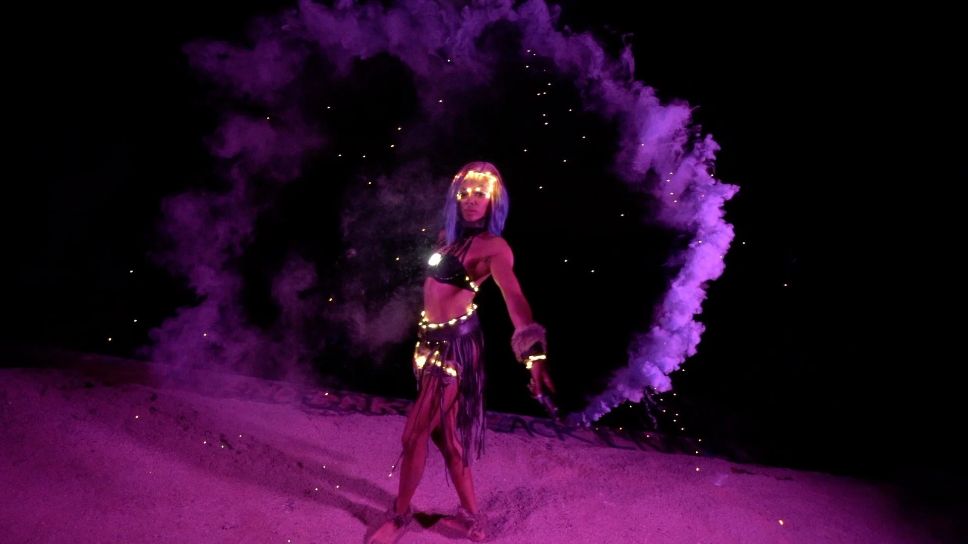 A woman with blonde hair and a choker, dressed in a black bra and a fringed skirt, stands on a sandy surface at night. She is holding a smoke flare emitting purple smoke, creating a heart-shaped formation in the dark sky behind her, illuminated by small sparks.