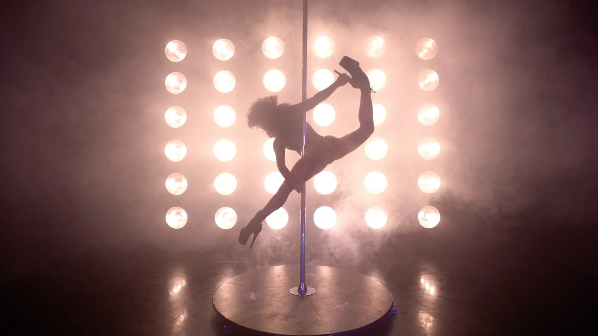 Silhouette of a woman performing a pole dance on a stage with bright lights in the background.