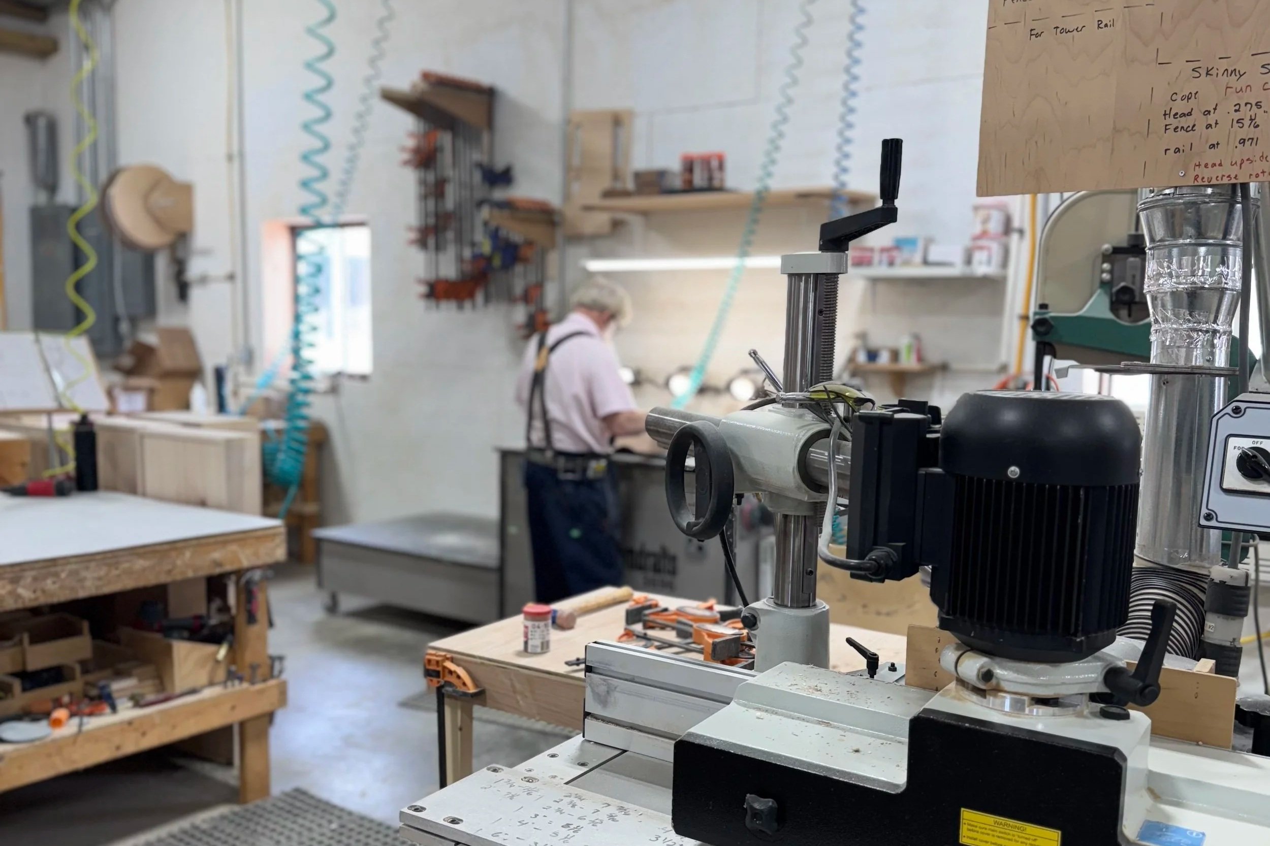 Close-up of a machine tool in a woodworking shop, with a blurred background of a person working at a workbench and shelves with tools and materials.
