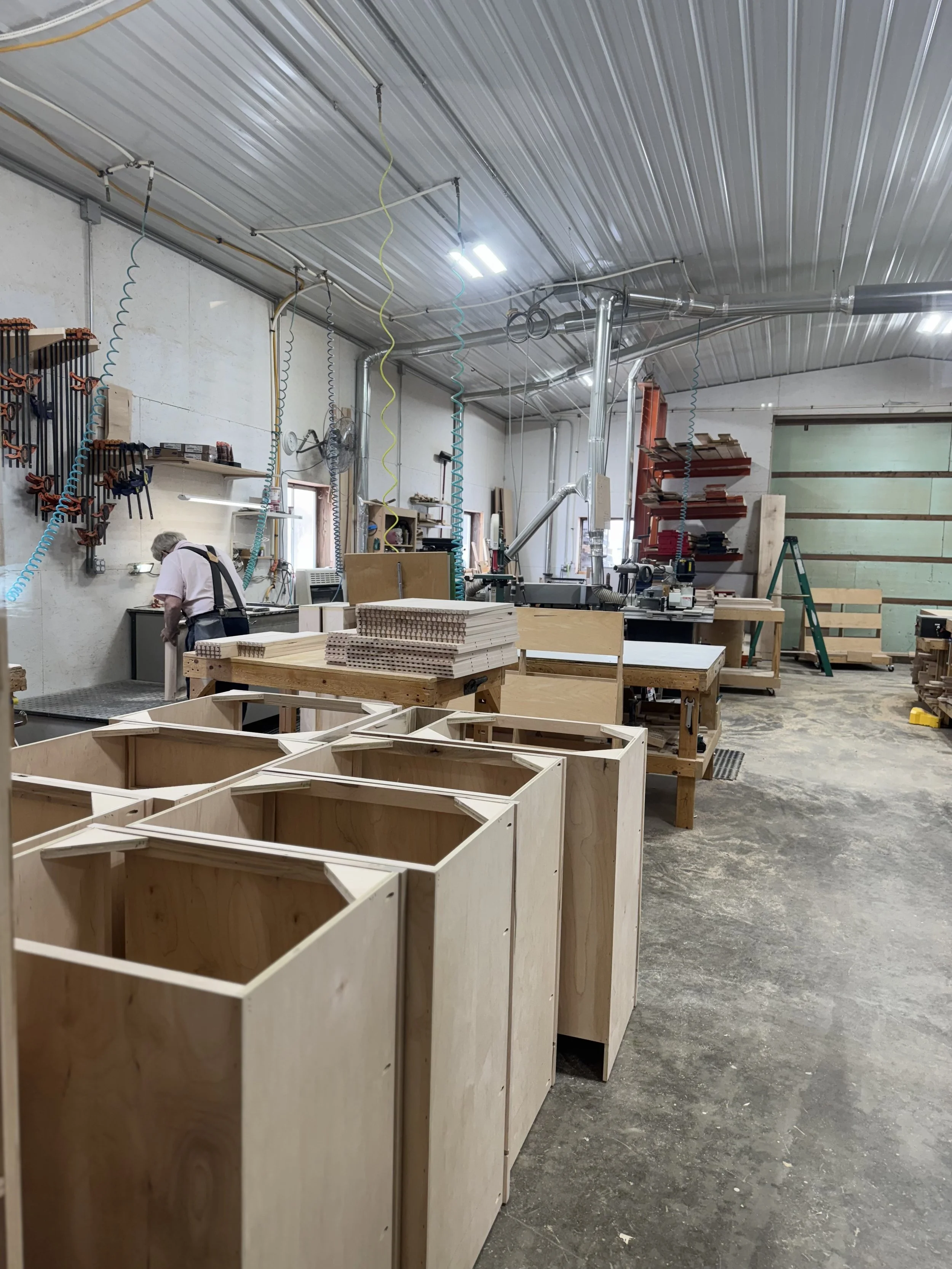 Woodworking workshop with unfinished wooden cabinets, tools, and materials, worker focused on project, shelves with clamps, and equipment for woodworking.