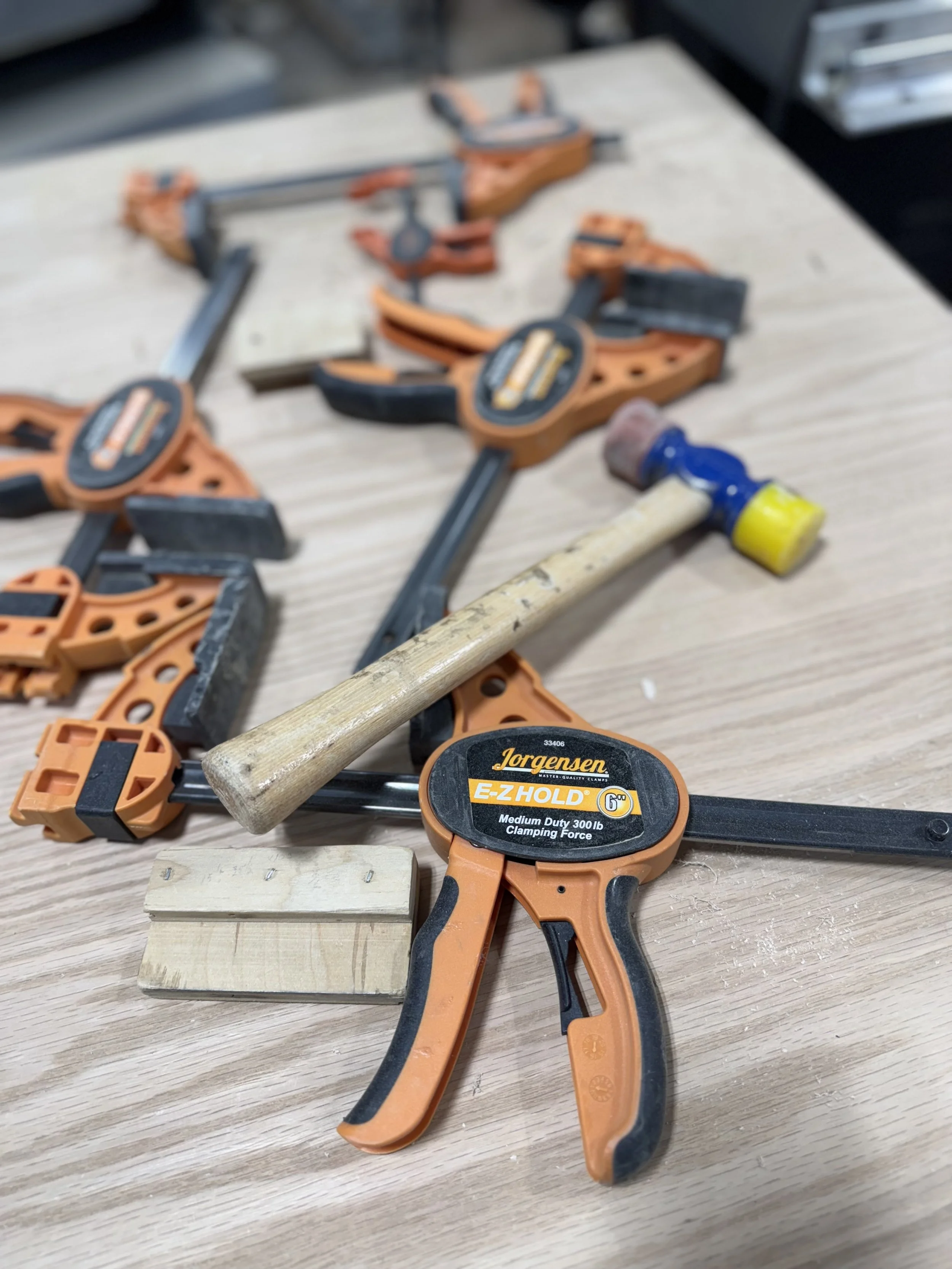 Clamps, hammer, and piece of wood on a wooden workbench.