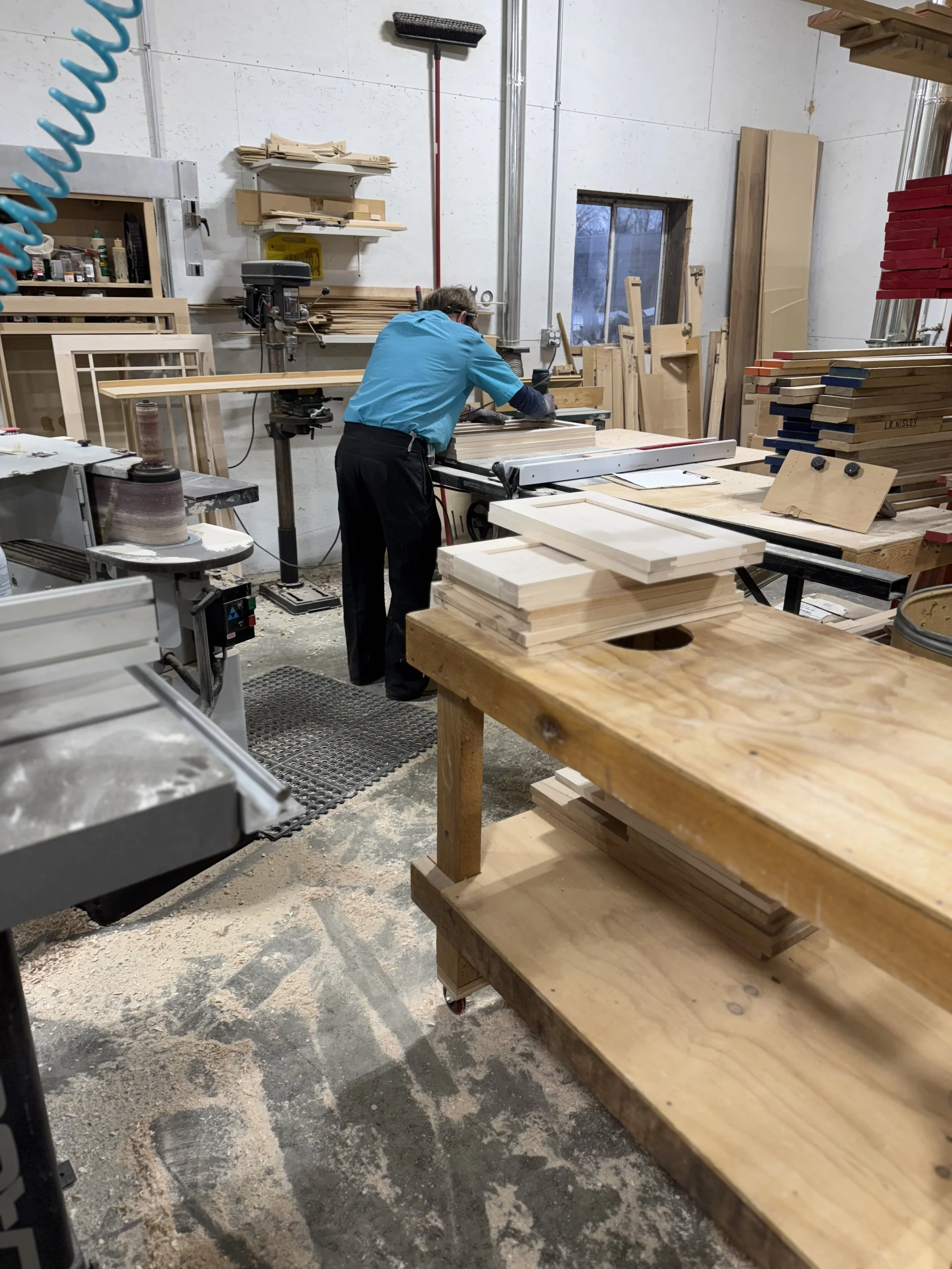A person working in a woodworking shop, cutting or shaping wood at a workbench, surrounded by shelves with wood pieces and tools.