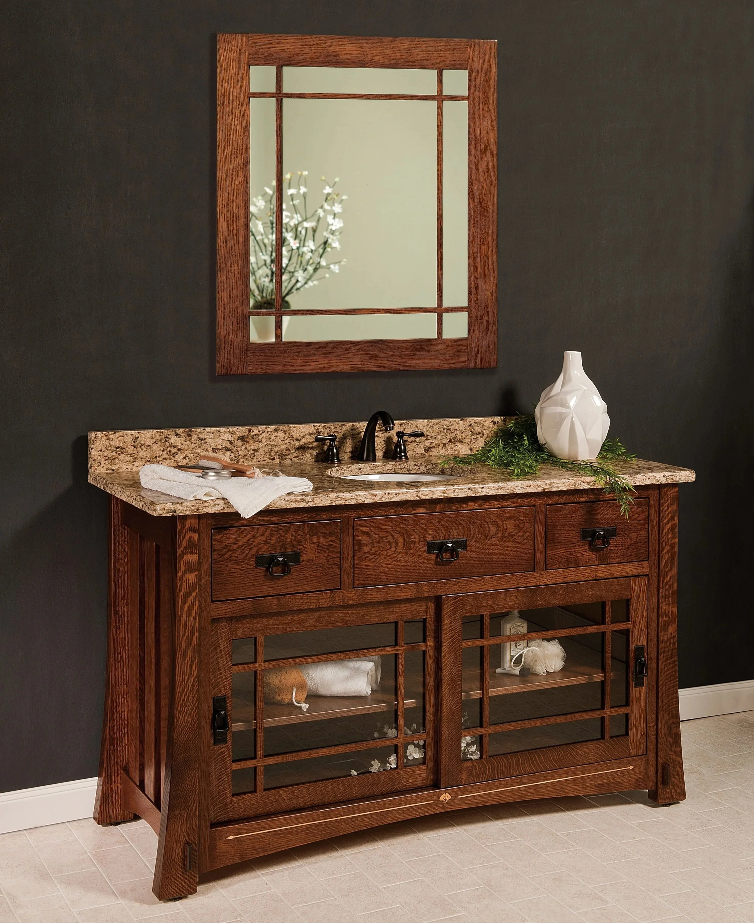 A wooden bathroom vanity with a granite countertop, a black faucet, and a mirror above. The vanity has glass-paneled cabinet doors, a white vase with greenery, and a rolled towel inside.