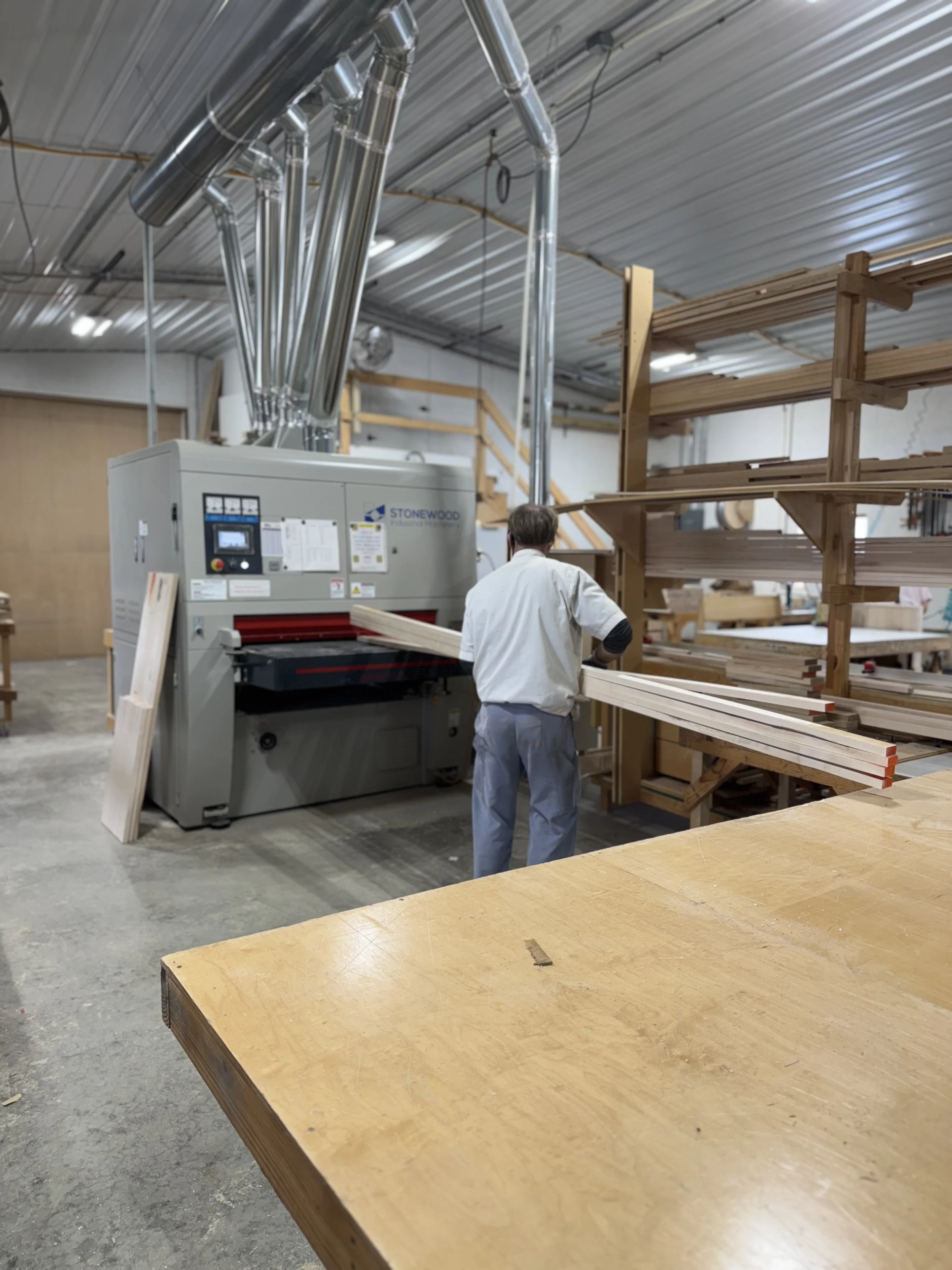 A woodworker in a workshop operating a large industrial wood processing machine, with shelves of cut wood and a wooden worktable in the background.