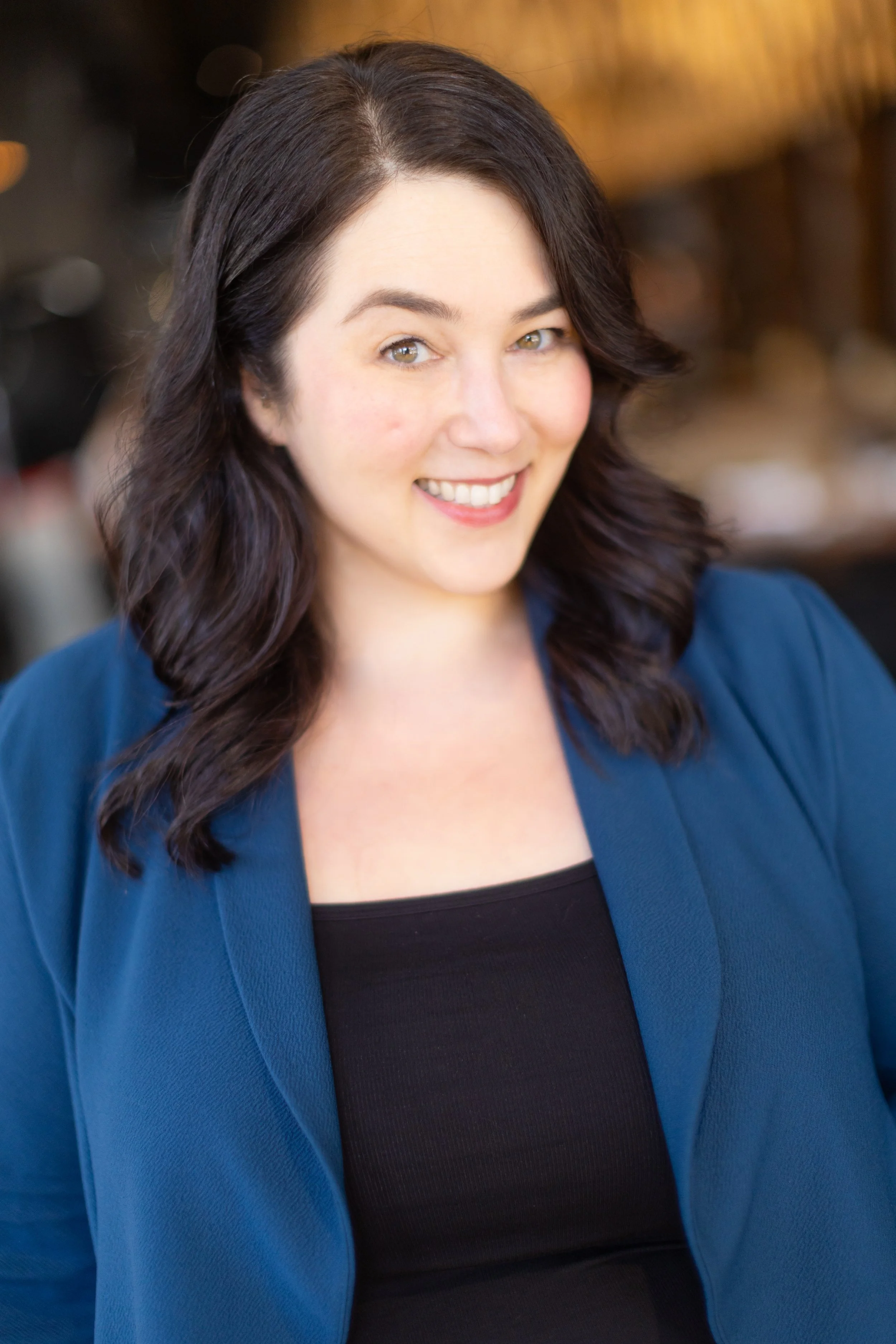 A woman with shoulder-length dark hair wearing a blue blazer and black top, smiling at the camera, with a blurred background.