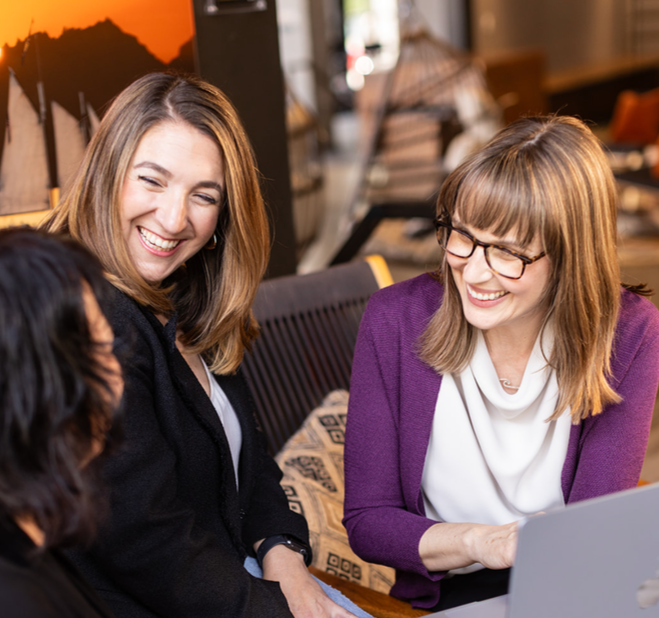 Three women sitting together at a table in a cafe or restaurant, smiling and laughing as they look at each other and a laptop.