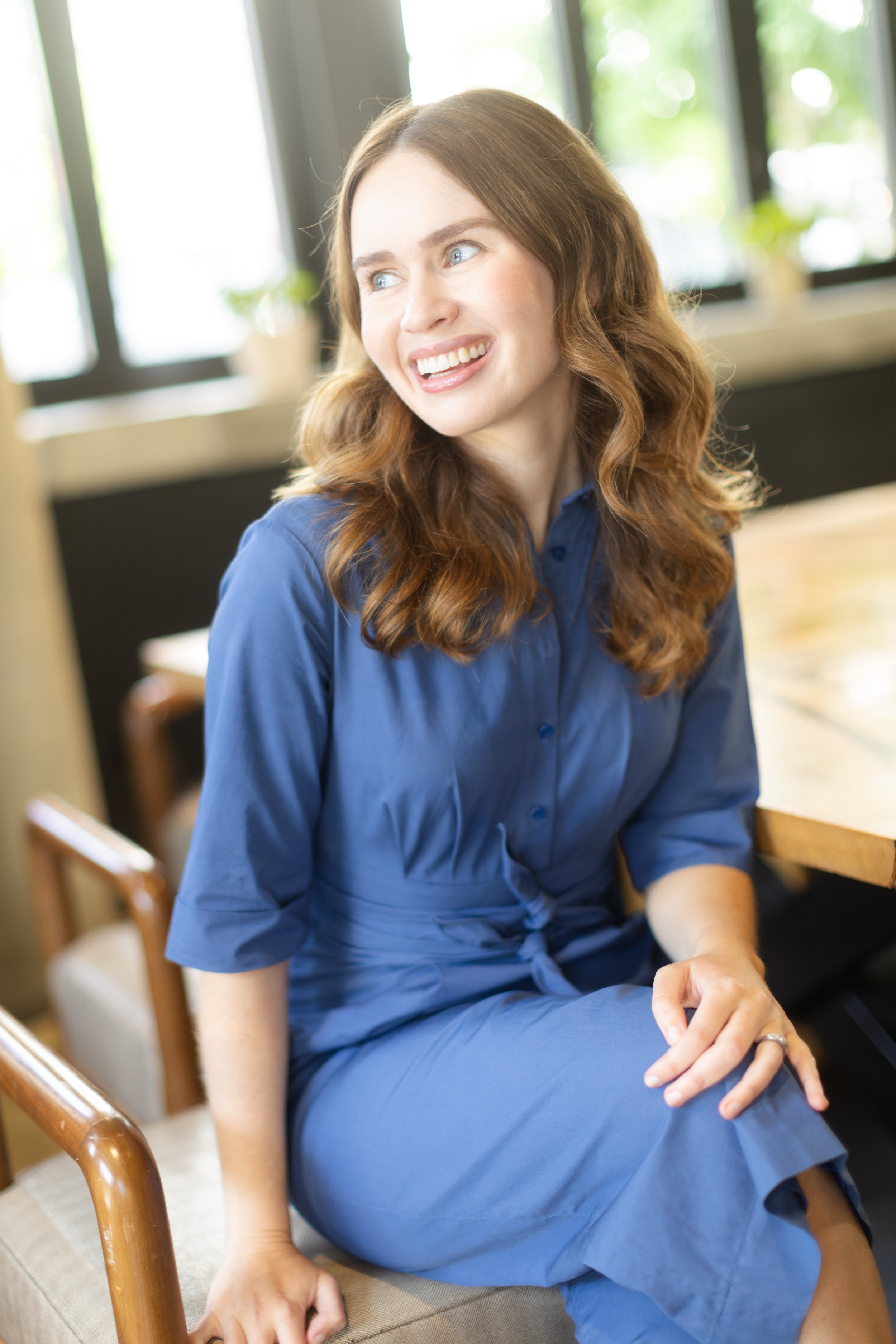 A woman with wavy, red hair wearing a blue dress, sitting on a chair in a well-lit room with large windows and plants in the background.