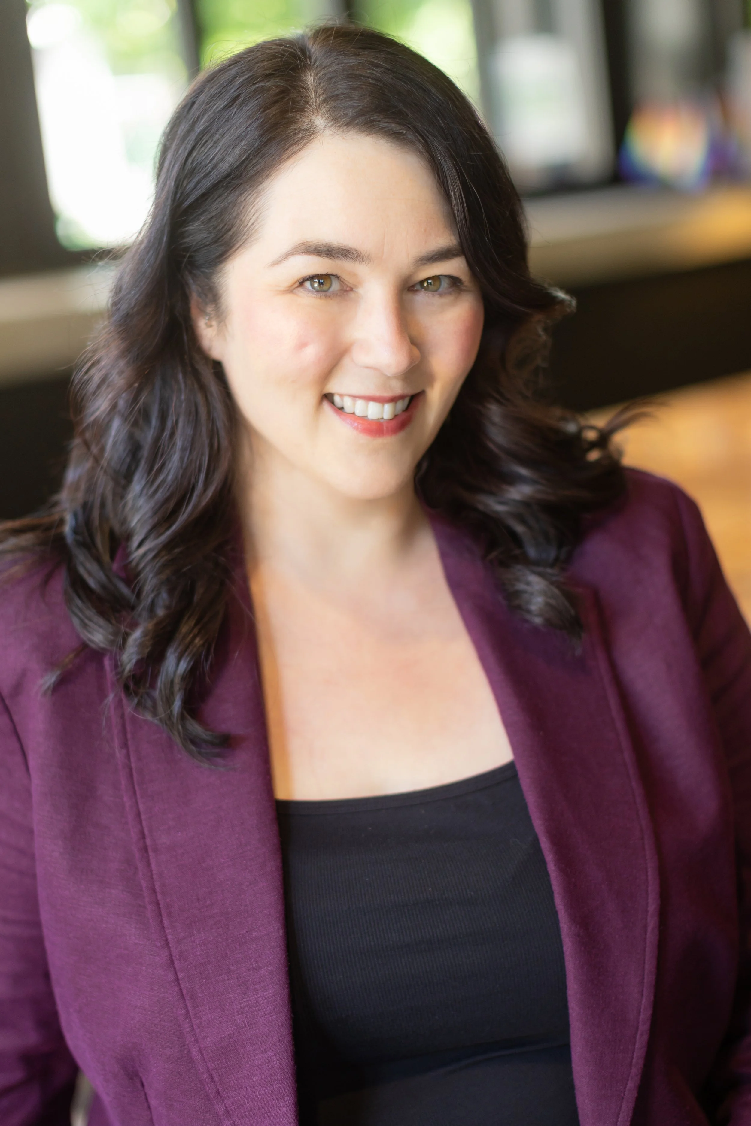 A woman with dark brown wavy hair smiling, wearing a black top and burgundy blazer, in a well-lit room with a blurred background.
