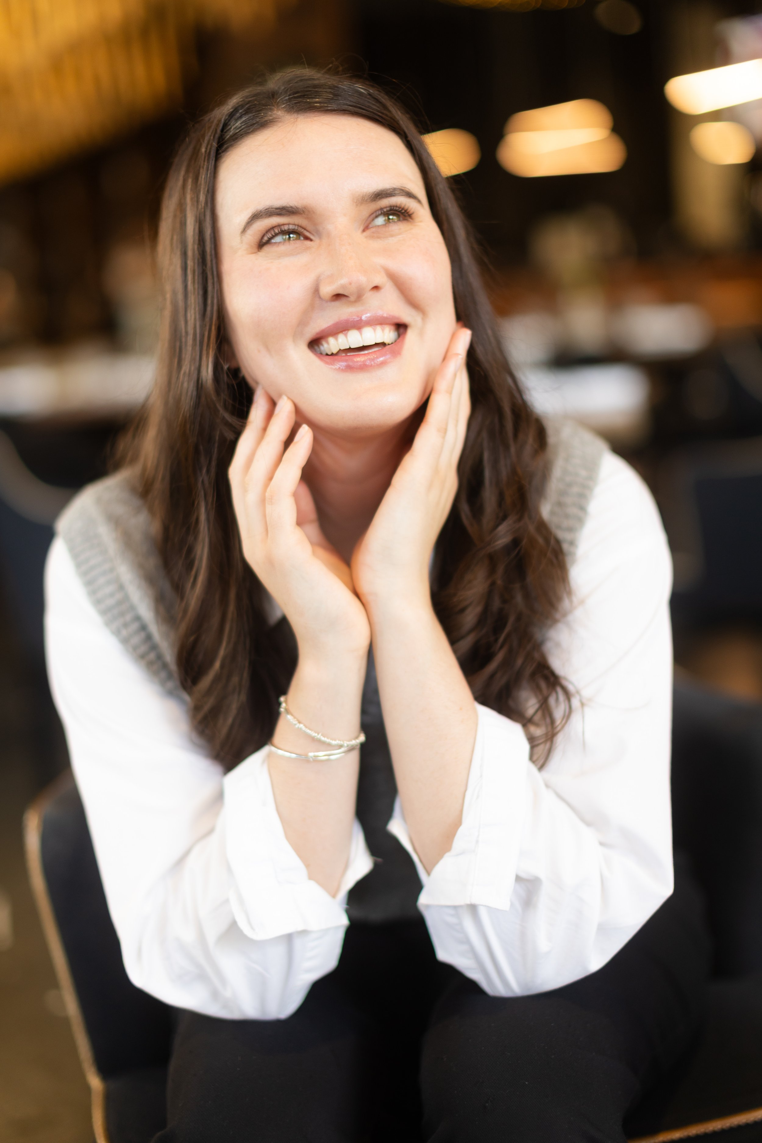 A young woman with long dark hair, smiling, holding her face with both hands, sitting indoors with a blurred background.