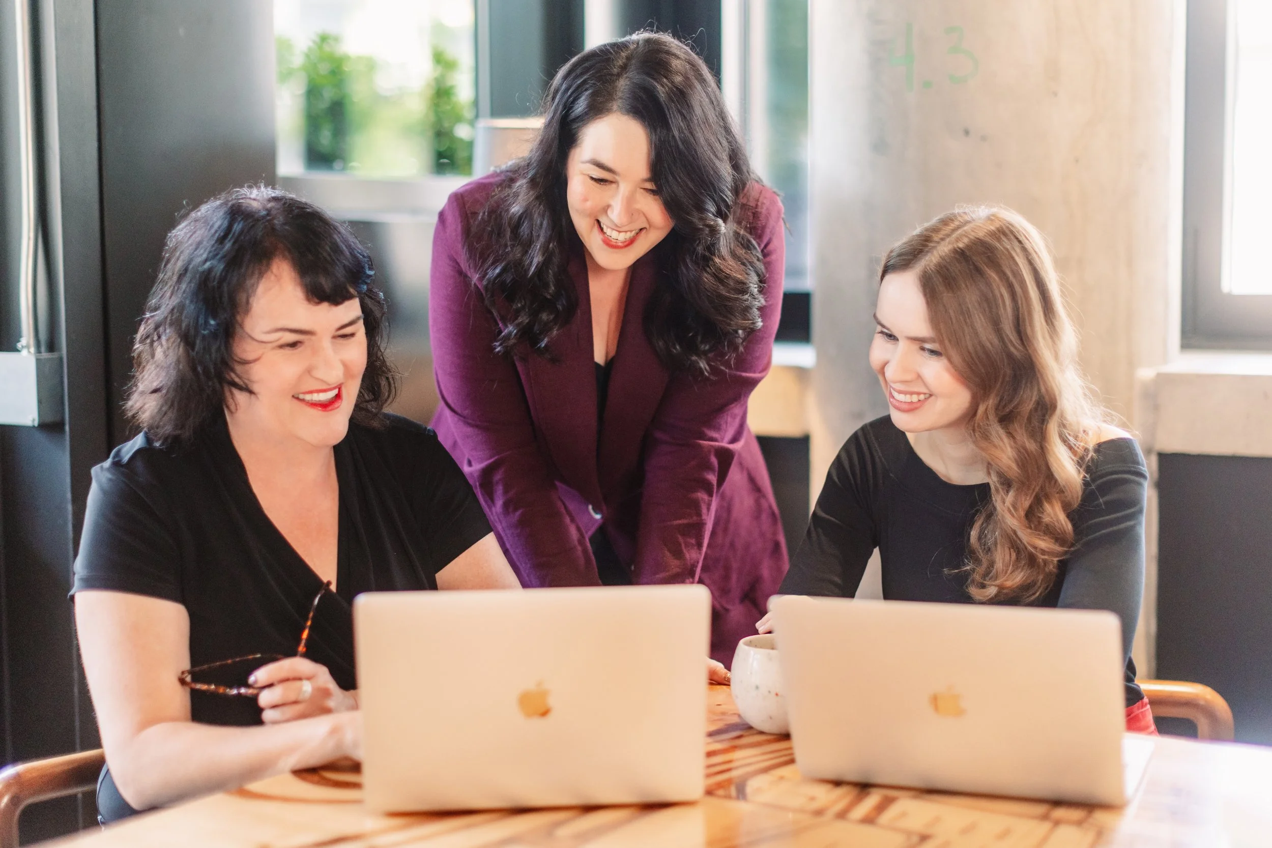 Three women smiling and looking at laptops on a wooden table in a bright room with large windows.