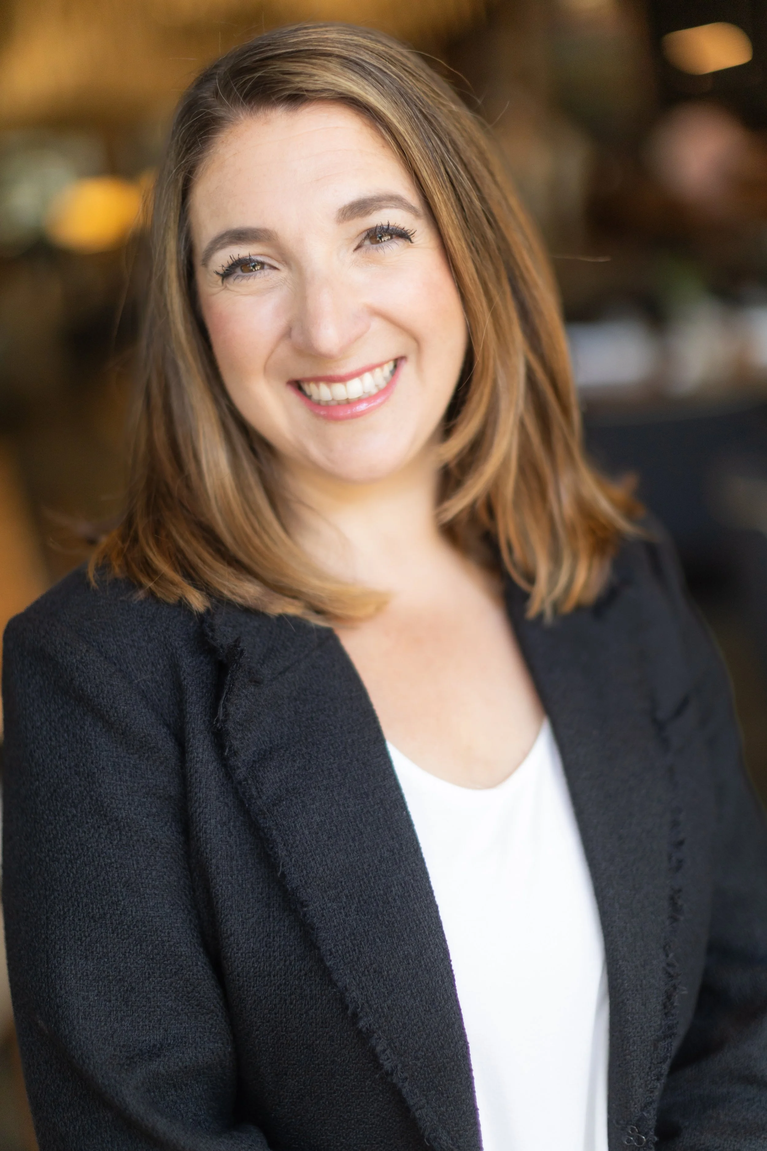 A smiling woman with shoulder-length brown hair, wearing a black blazer over a white top, in an indoor setting.