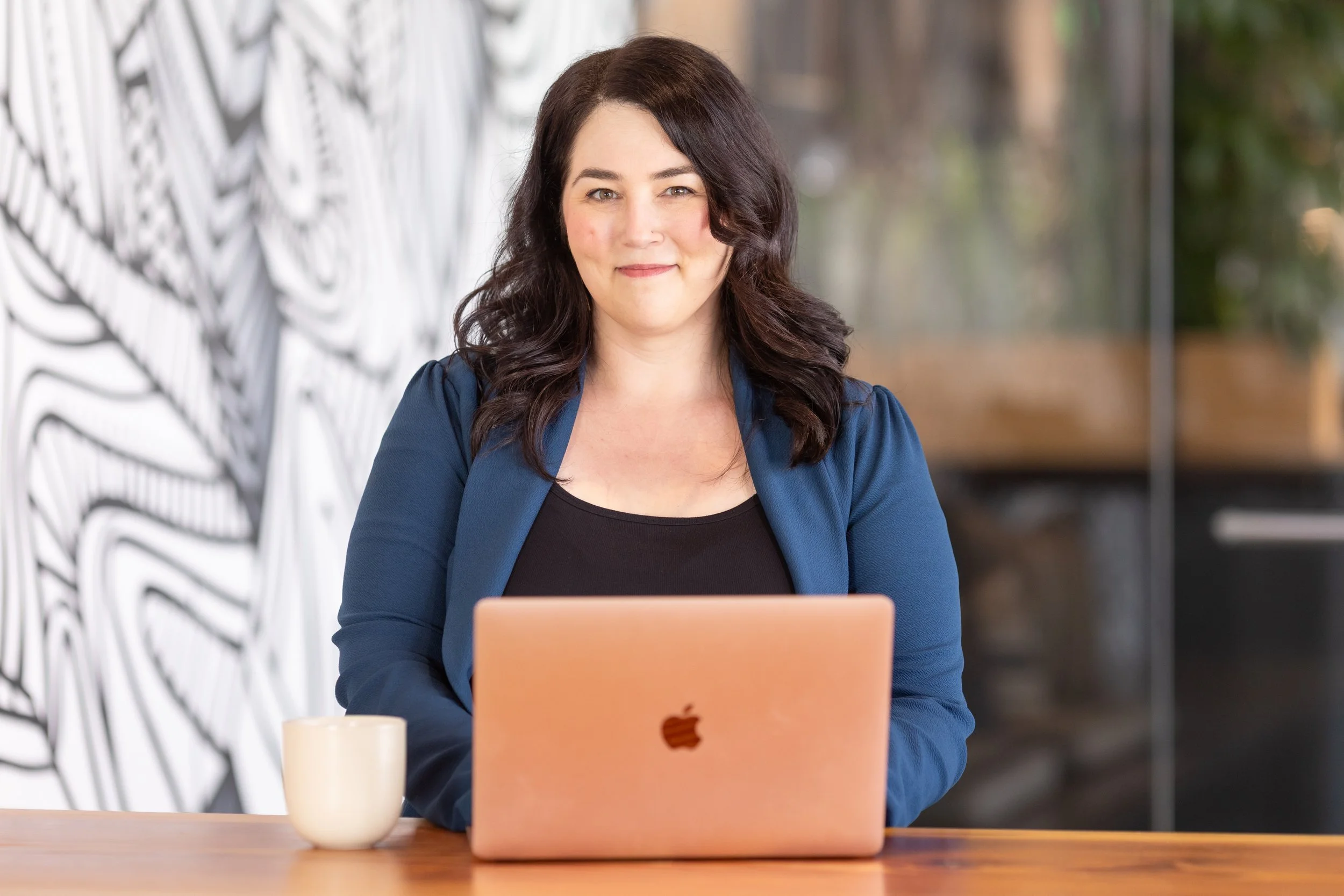A woman with dark hair sitting at a wooden table using a pink Apple MacBook, with a coffee mug beside her, in a modern cafe or office environment.
