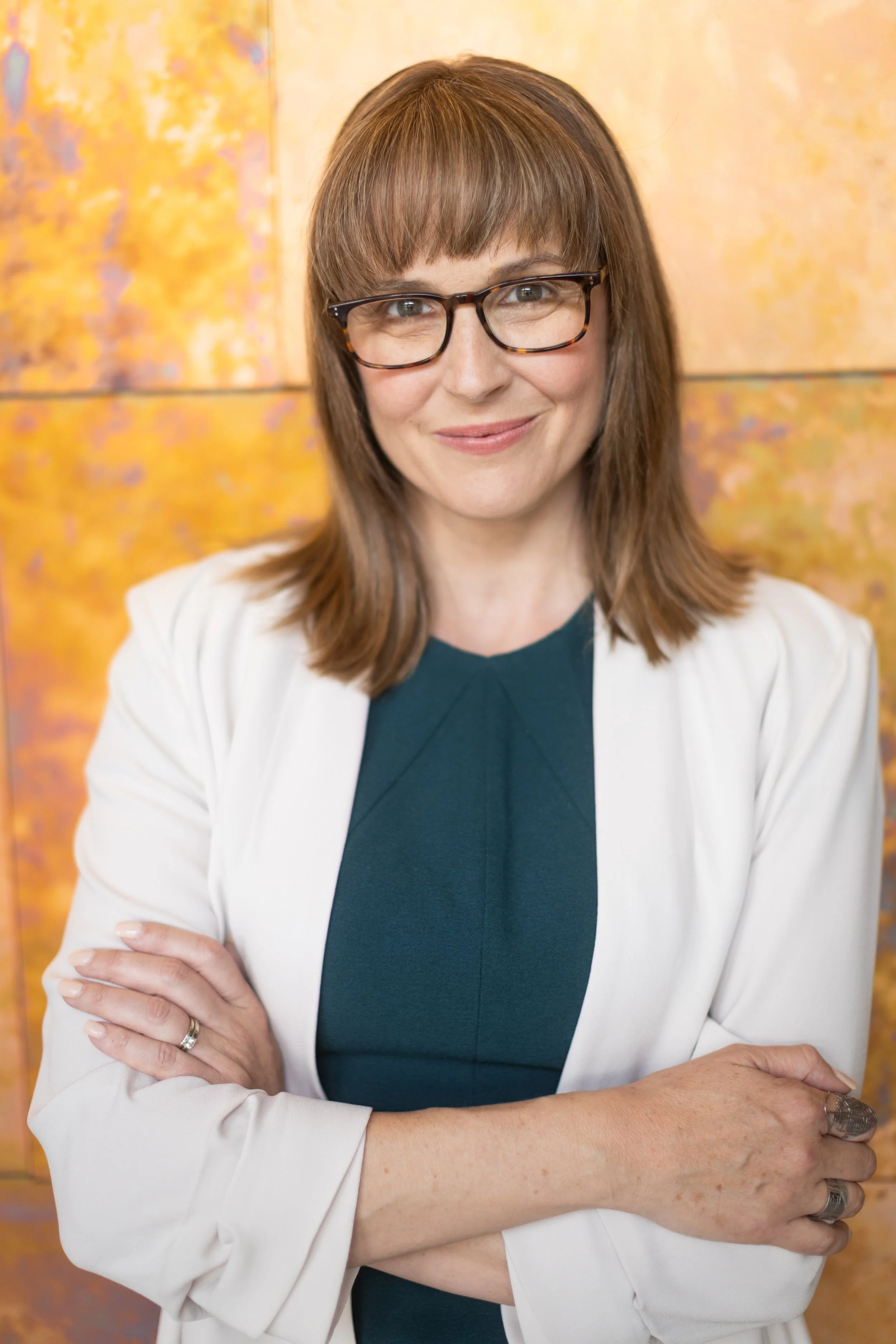 A woman with shoulder-length brown hair and glasses smiling, crossing her arms, wearing a white blazer over a dark top, standing against a colorful, abstract background.