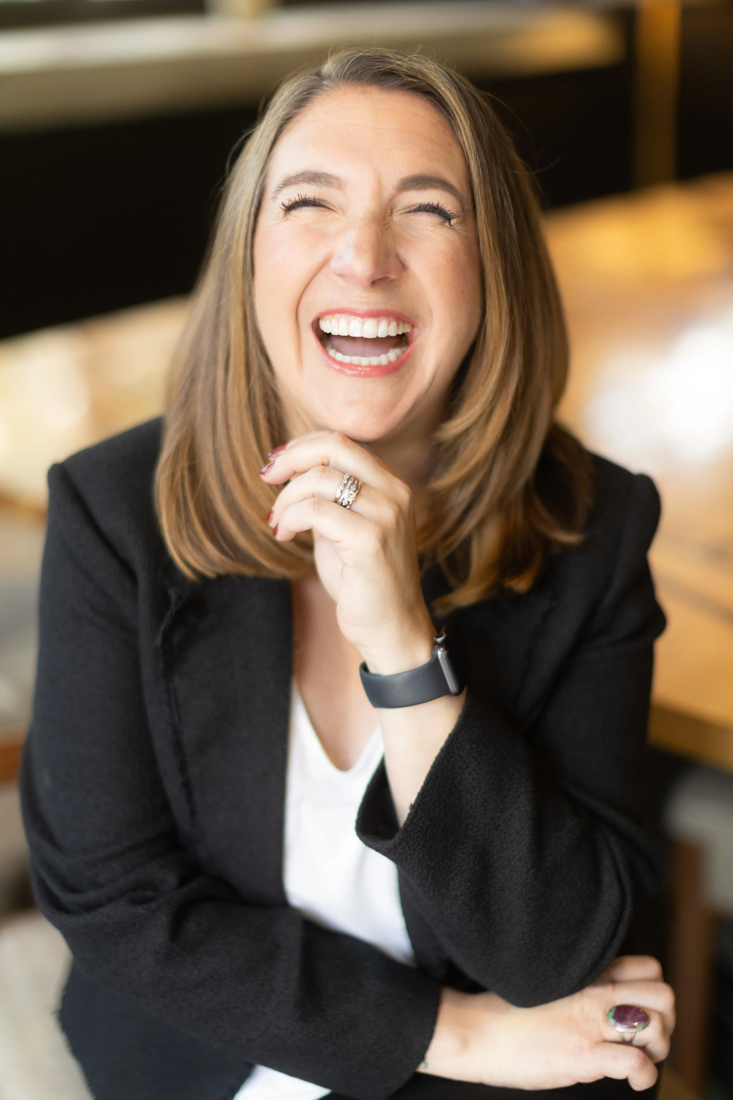 Woman laughing, wearing a black blazer and a white shirt, with a smartwatch and rings, sitting with her hand near her chin.