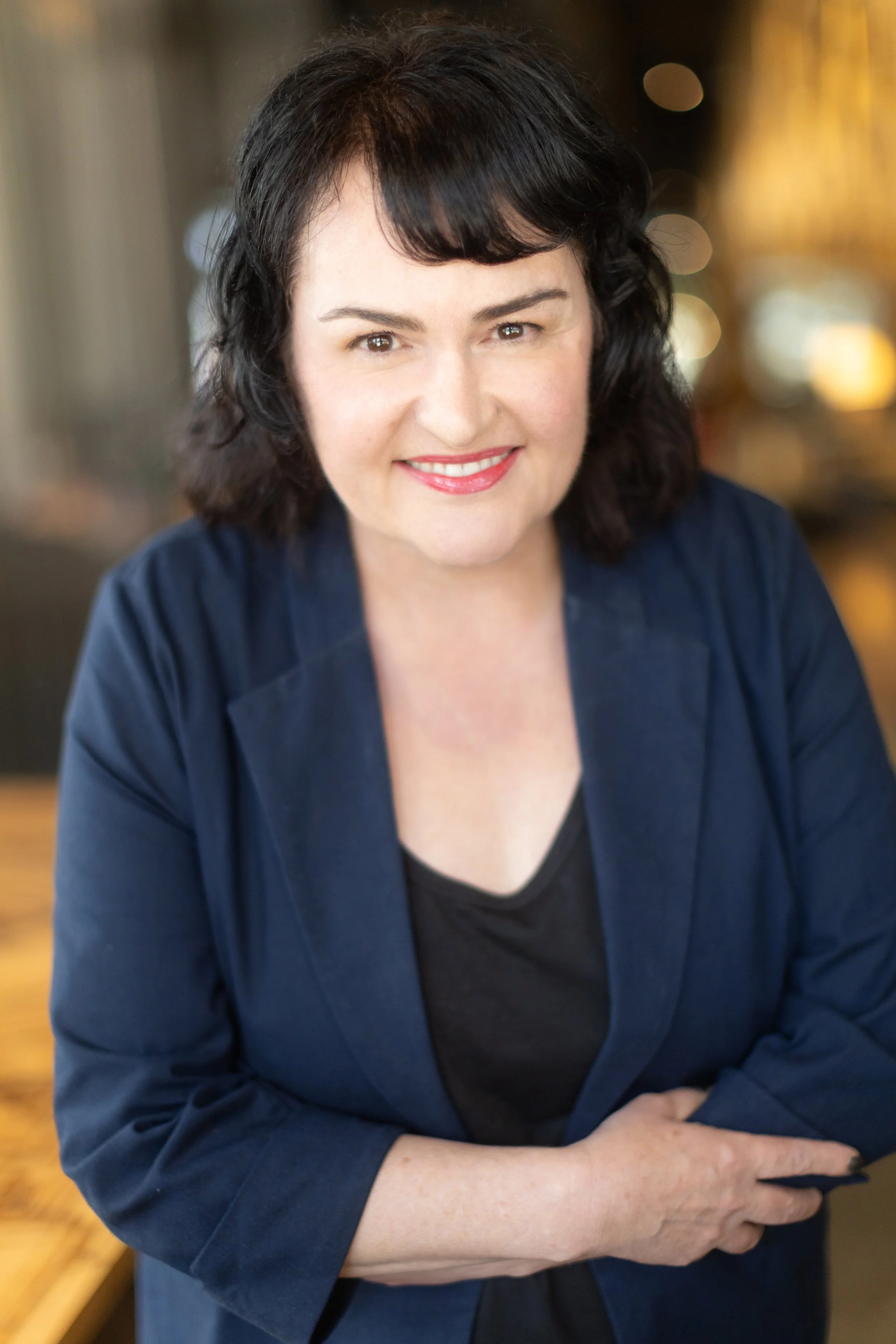 A woman with dark, shoulder-length hair, smiling, wearing a dark blue blazer and black top, sitting in a warmly lit indoor setting.