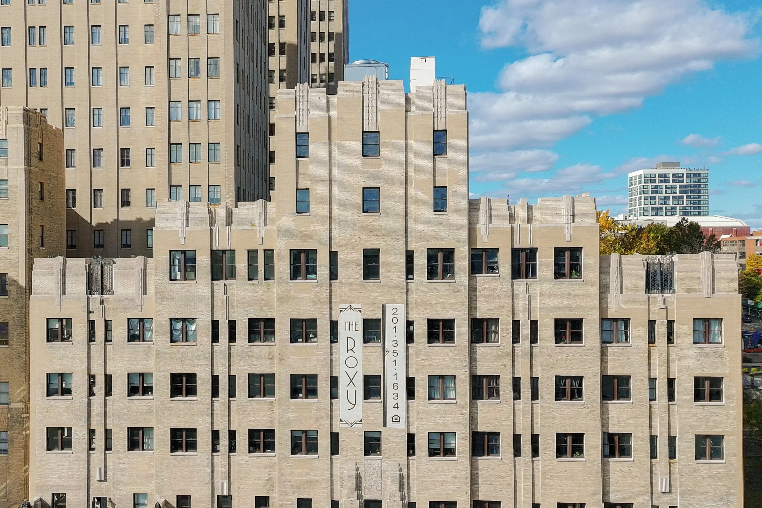Front view of a beige brick apartment building, The Roxy in an urban setting under a blue sky with scattered clouds.