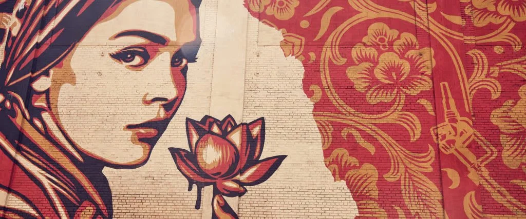 Wall mural of a woman's face with a lotus flower in front of her, painted in a stylized, graphic art style, on a brick building with floral patterns and a pointing hand in the background.