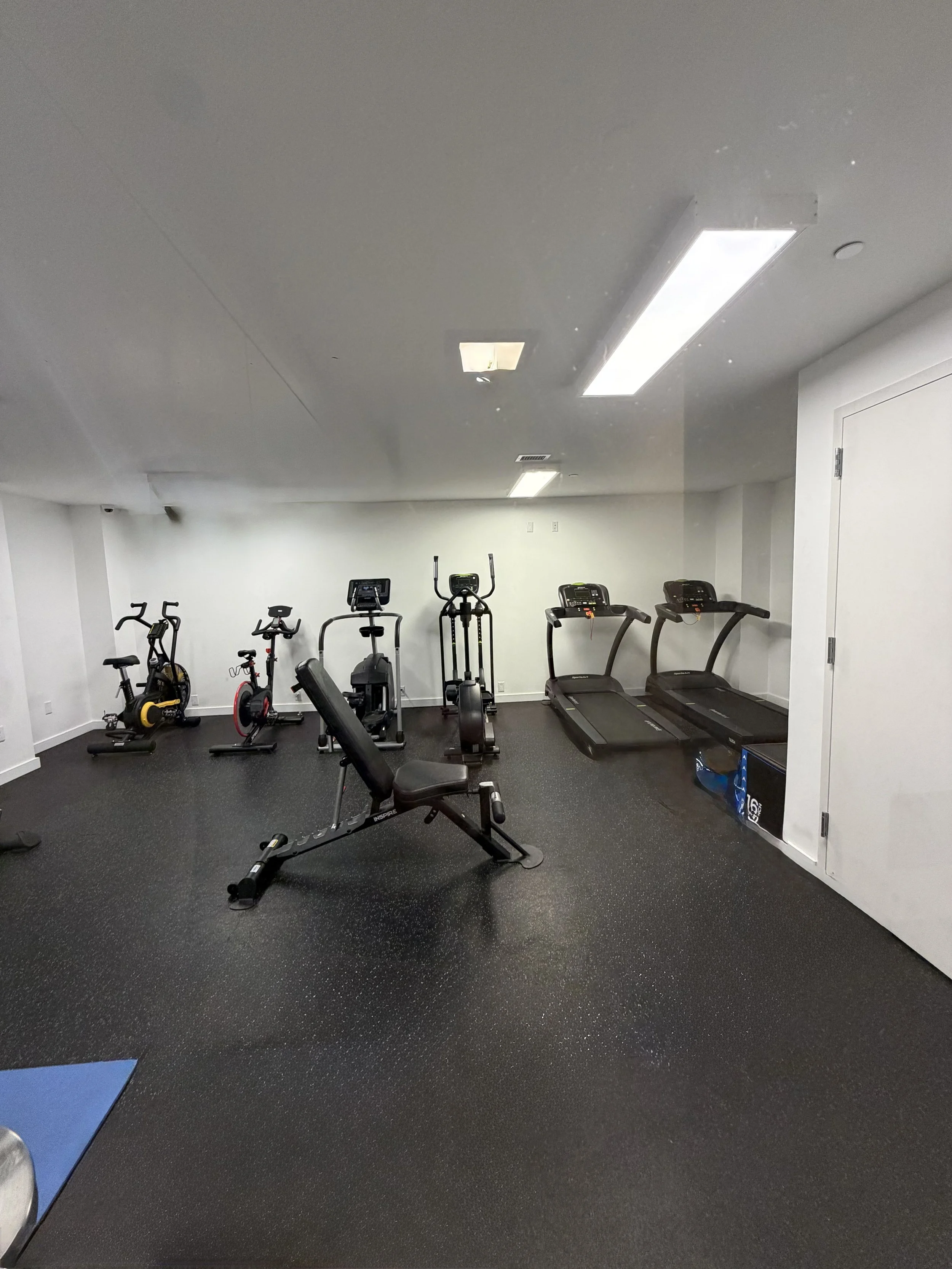 Empty gym room with exercise equipment including stationary bikes, ellipticals, treadmills, and a workout bench on black rubber flooring.