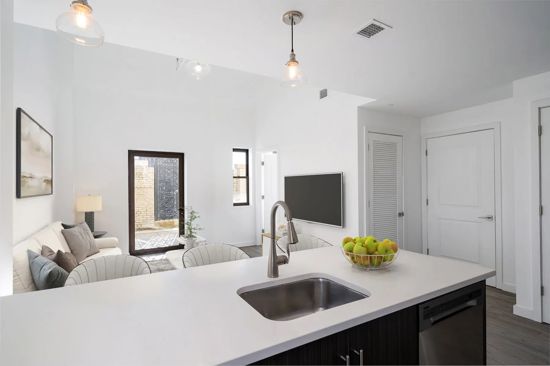 Modern kitchen with white counters, a bowl of green apples, and a view of the living room with a sofa, lamp, and TV.