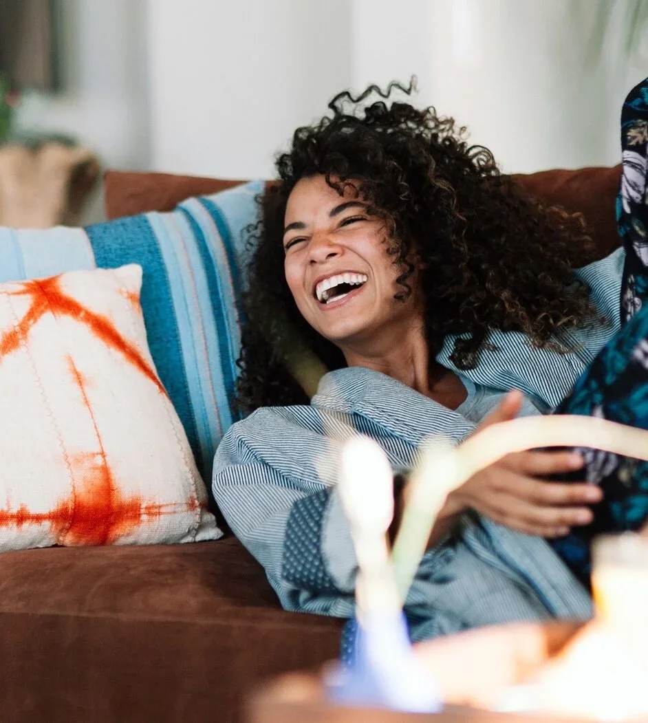 A woman with curly hair laughing while sitting on a couch. She is wearing a light blue striped shirt and is next to a pillow with orange and white tie-dye patterns.