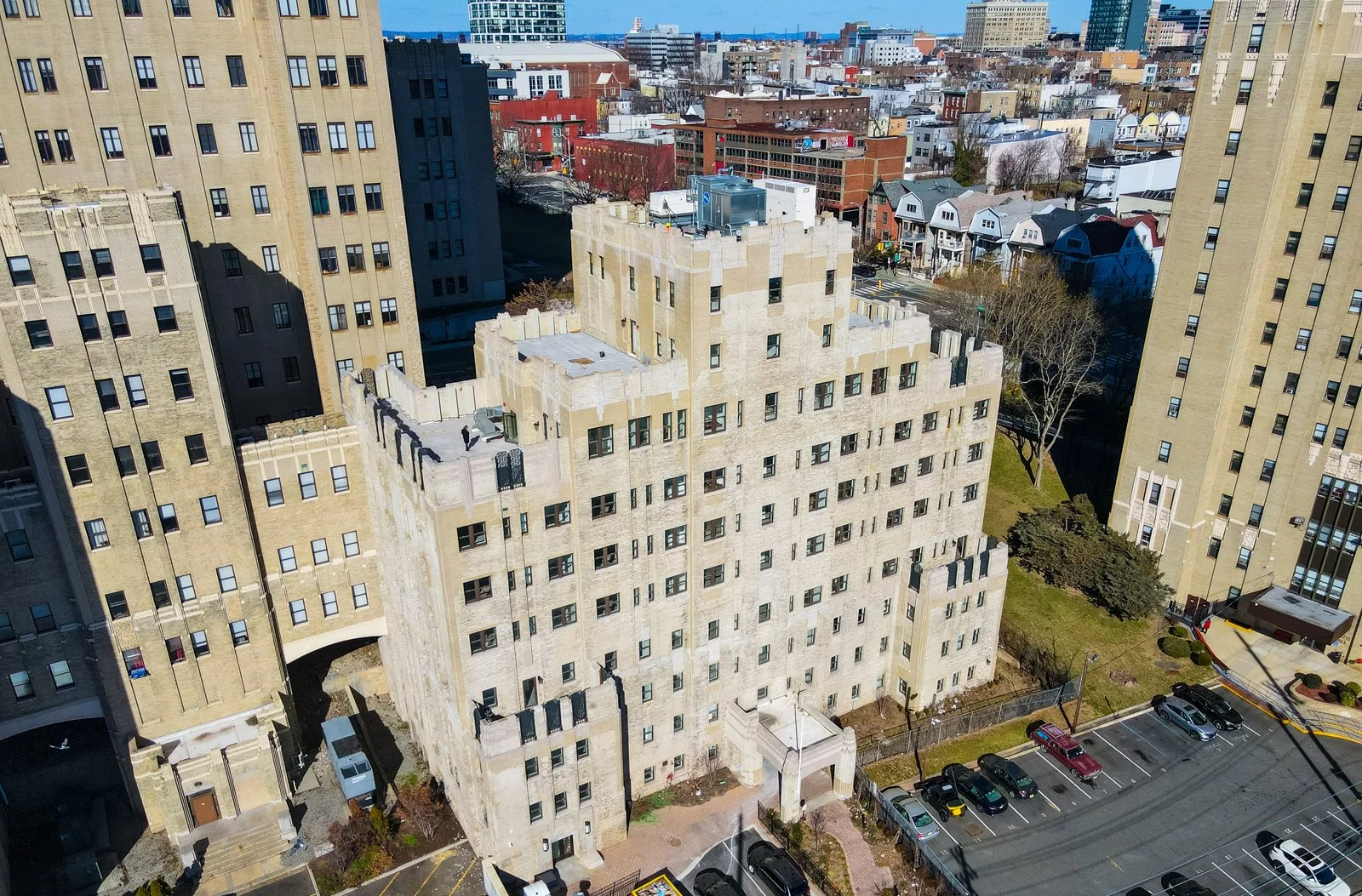 An aerial view of a beige, multi-story building with a rooftop deck, surrounded by taller high-rise buildings and a parking lot with several parked cars.